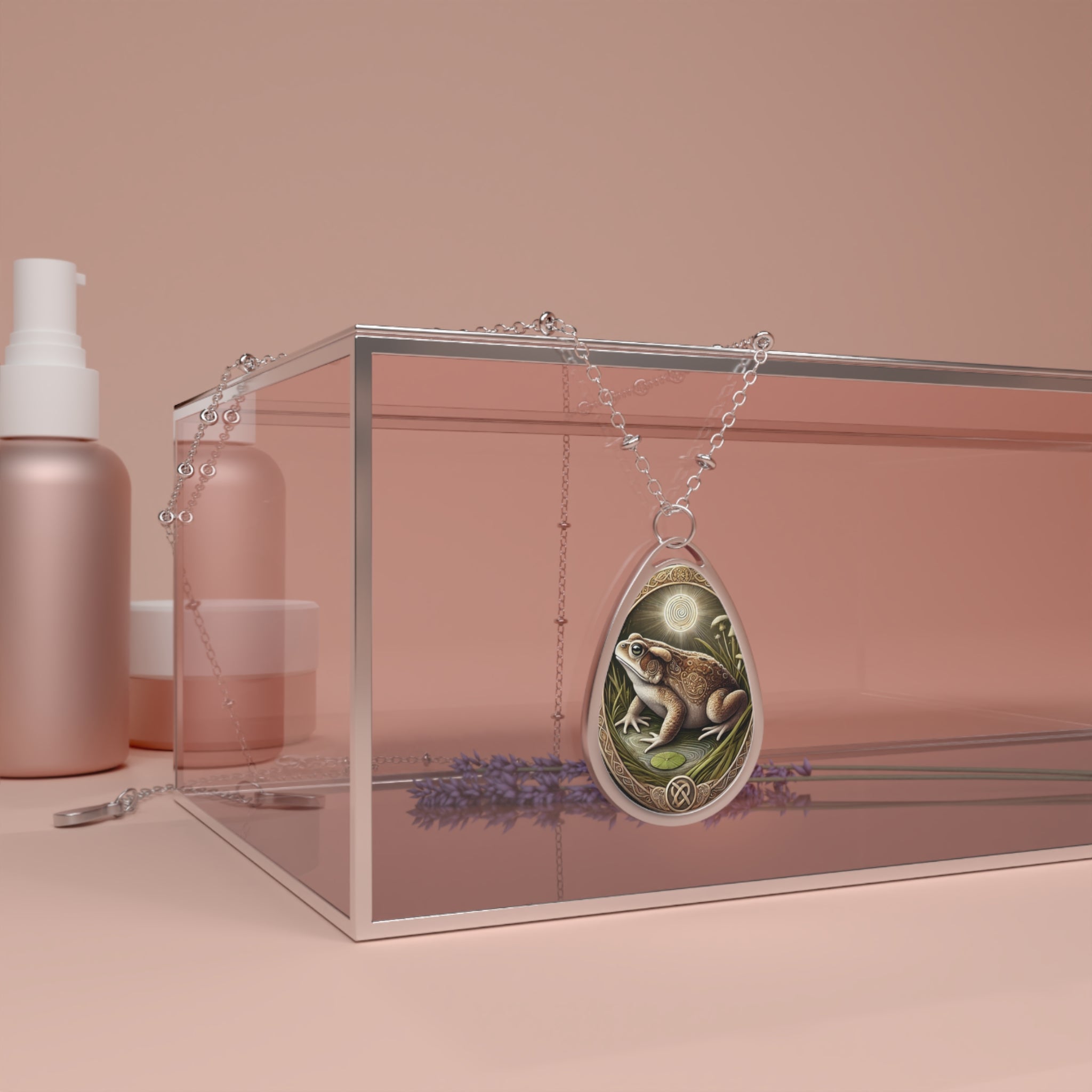 A silver necklace featuring a toad pendant draped over a clear box with lavender sprigs and cosmetics in the background, set against a soft pink backdrop. The pendant displays a detailed toad in a natural setting with lush greenery and a mystical spiral design above, symbolizing transformation and grounding.