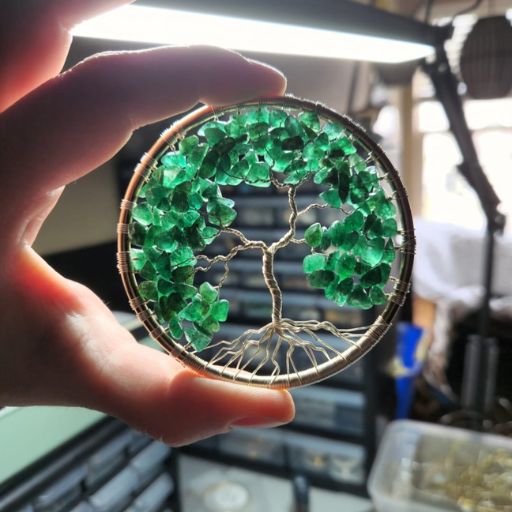 A hand holding a circular wire-wrapped pendant with an intricate tree design, adorned with numerous small green crystals, backlit by a bright light source, with a blurred background of a workspace filled with tools.