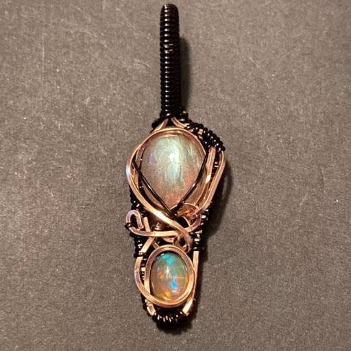 An intricate wire-wrapped pendant displaying two iridescent opal stones with pink and green hues, nested within a dark copper wire design on a black background.