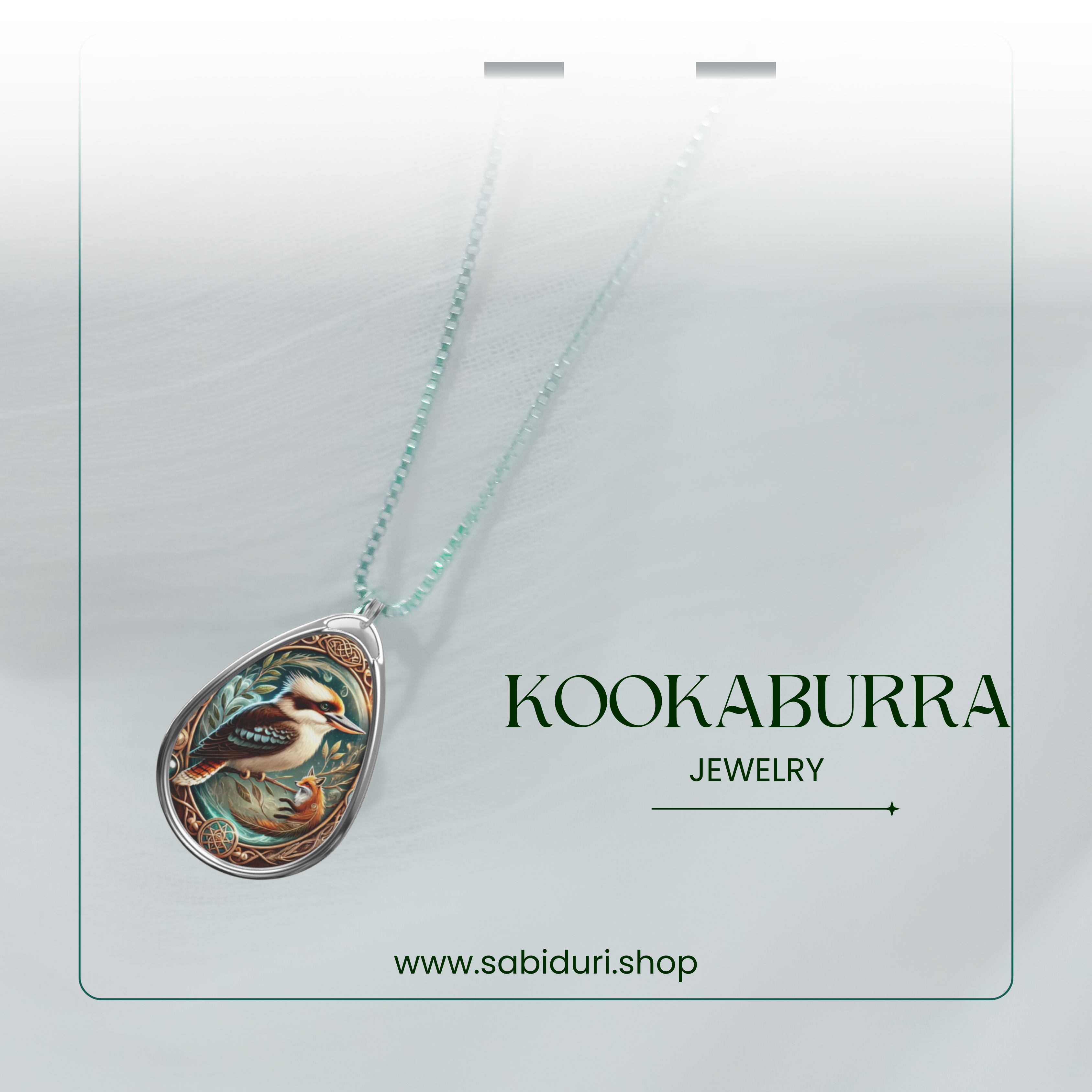 A glowing kookaburra pendant is suspended from a silver chain, displayed against a soft, misty white background. The intricate illustration of the kookaburra is accentuated by swirling gold details and blue-green botanical elements. The text "KOOKABURRA" and "JEWELRY" appears, along with "www.sabiduri.shop."