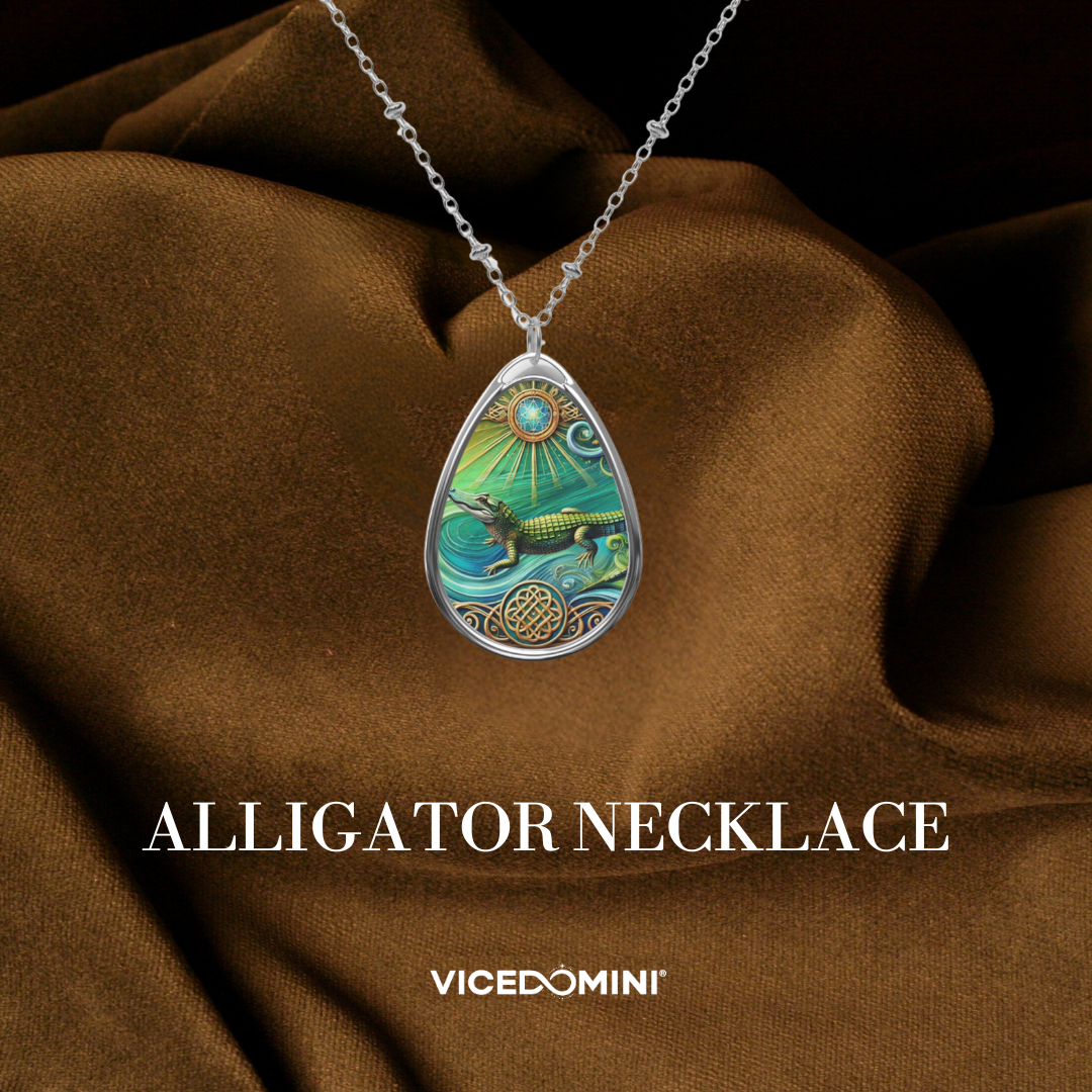 A teardrop-shaped silver pendant necklace featuring an intricate alligator illustration in green, gold, and blue tones, displayed on draped brown fabric with the text 'ALLIGATOR NECKLACE' and 'VICEDOMINI' below.