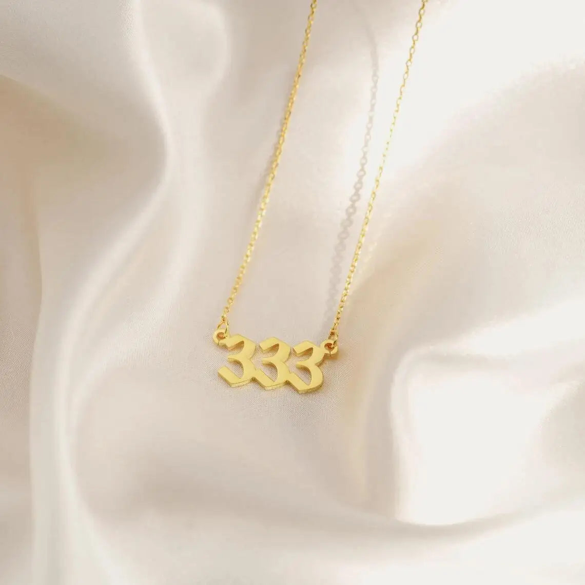 A gold angel number 333 necklace displayed on a smooth, white silk background. The pendant has a modern, bold font and is suspended from a delicate gold chain.