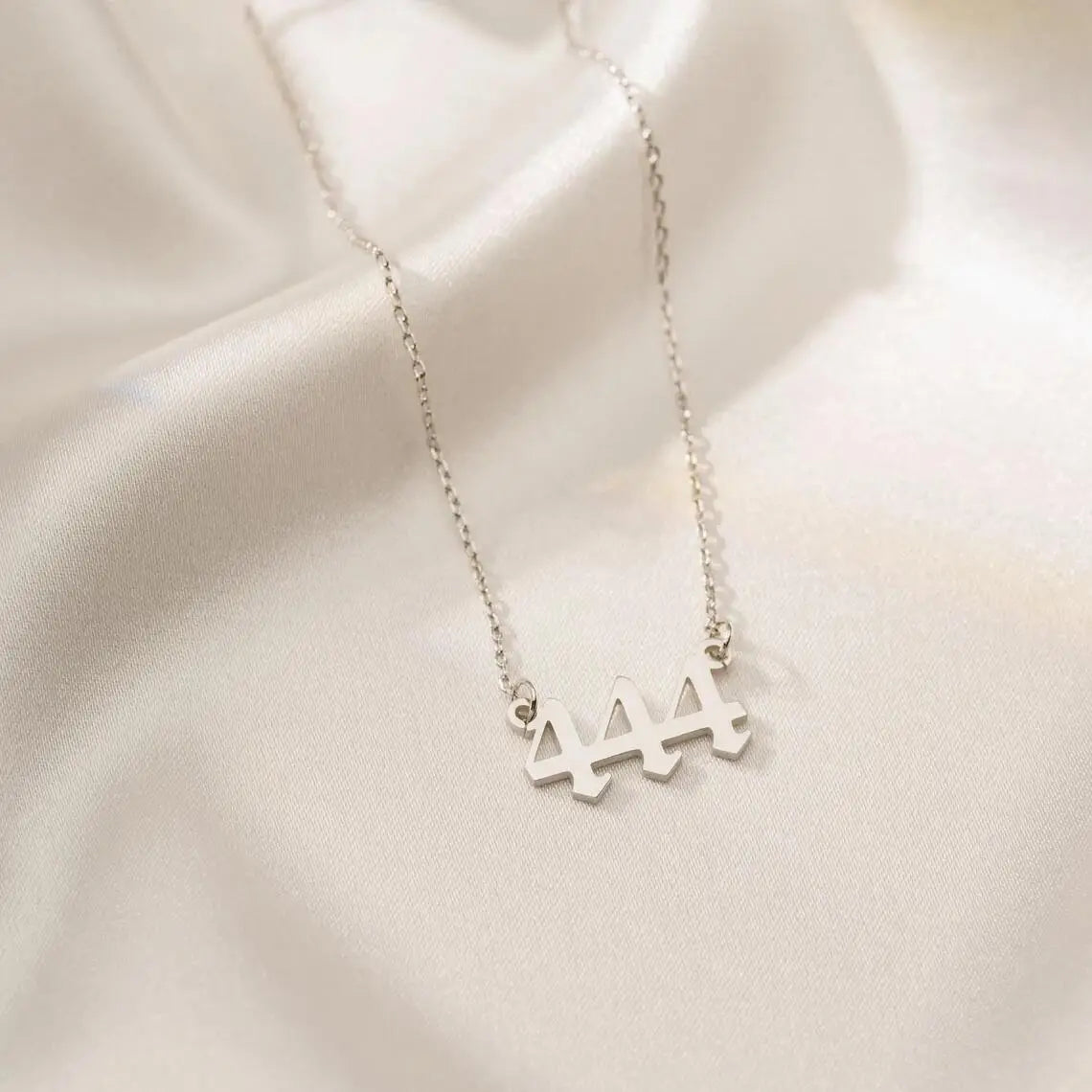 A close-up of a silver angel number 444 necklace resting on a soft, white silk background, emphasizing its polished finish and delicate chain.