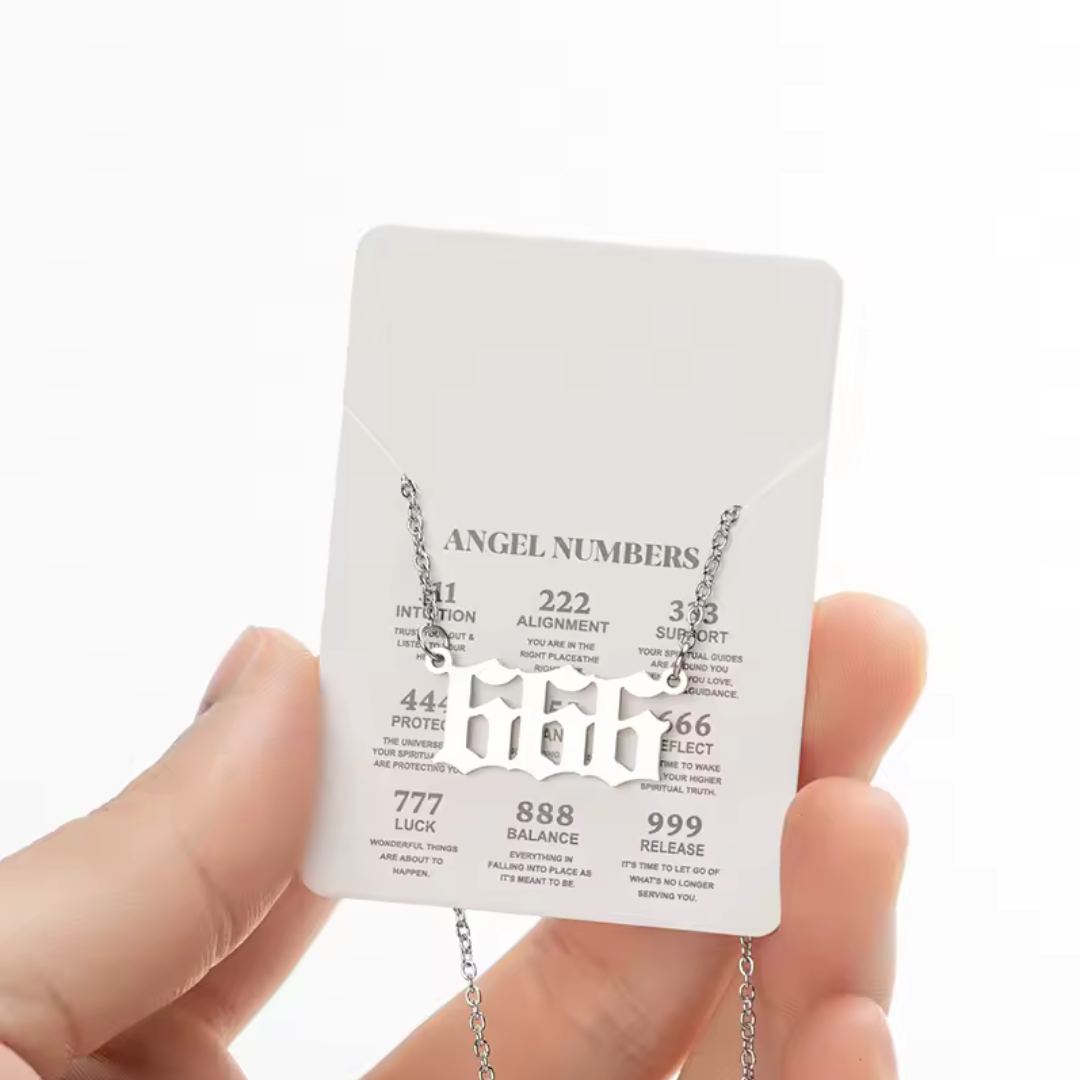 A silver angel number 666 necklace displayed on a white card listing various angel numbers and their meanings, such as intuition (111), alignment (222), and balance (888). The necklace is held by a hand, showc