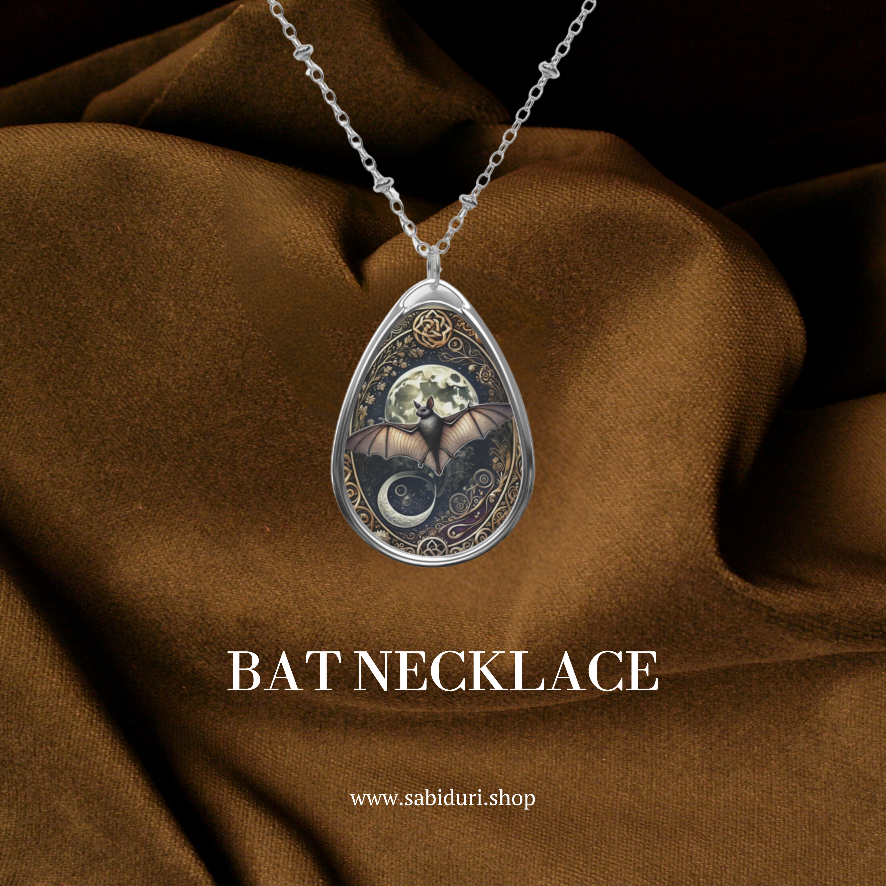 A silver bat necklace pendant with an intricately detailed bat and moon design, set against a rich brown fabric. The pendant combines celestial symbols and delicate scrollwork for a mystical vibe, with the text below reading "BAT Necklace" and the website "www.sabiduri.shop.