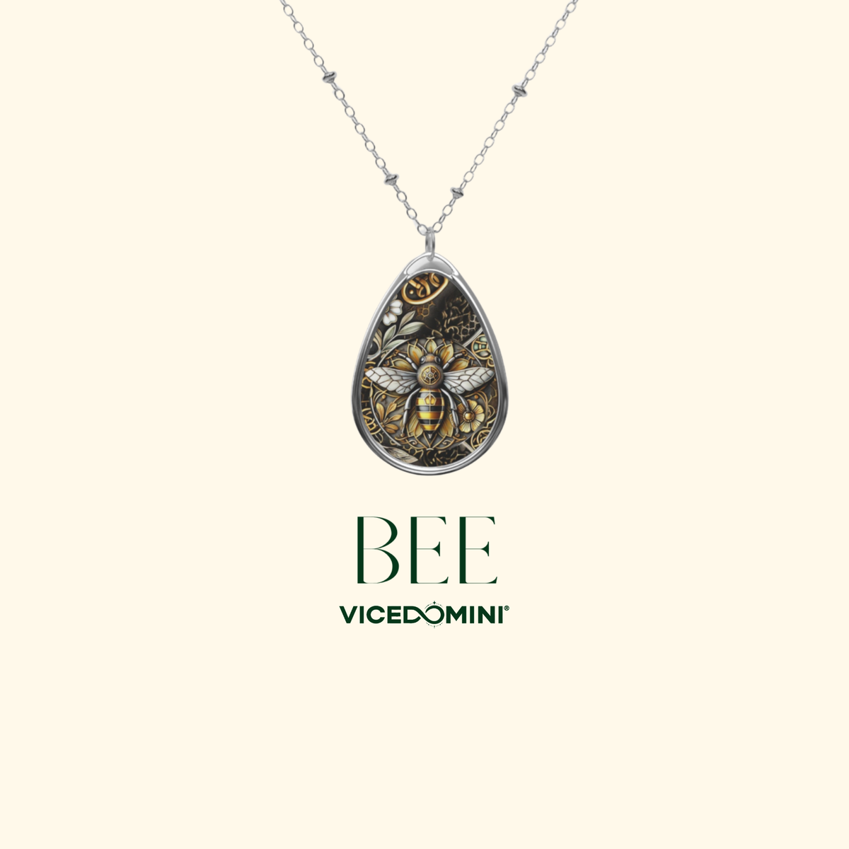 A teardrop-shaped pendant necklace featuring a detailed bee design in gold and silver tones, suspended from a delicate silver chain. The brand name 'VICEDOMINI' and the word 'BEE' are printed below the necklace on a solid cream background.