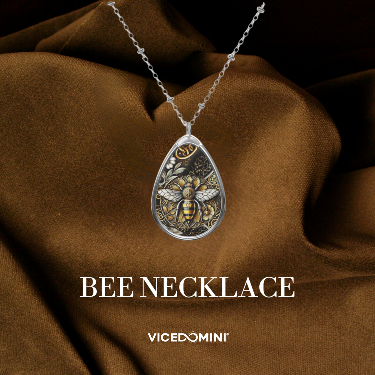 A teardrop-shaped silver pendant necklace featuring an intricate bee and floral design in gold and silver, draped over brown satin fabric with the text 'BEE NECKLACE' and 'VICEDOMINI' below.