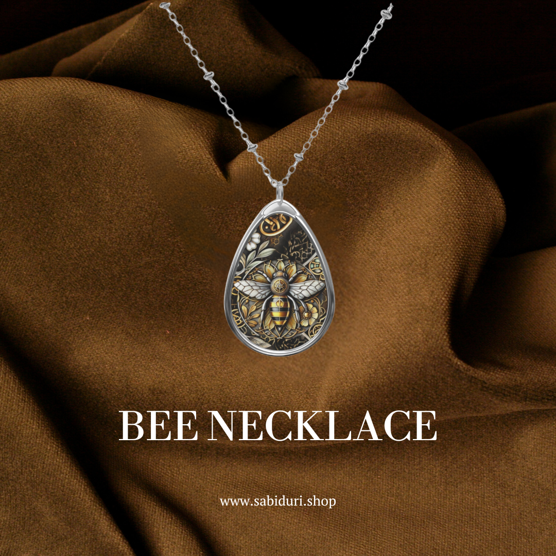 A close-up of the bee necklace, showcasing the detailed pendant design, resting on a rich brown fabric background. The text "BEE NECKLACE" is prominently displayed in white at the bottom, along with the URL "www.sabiduri.shop."