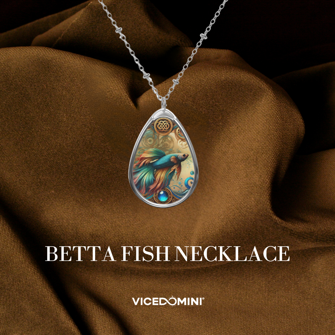 A teardrop-shaped pendant necklace featuring a colorful illustration of a betta fish, suspended from a silver chain and displayed against draped brown satin fabric. The text 'BETTA FISH NECKLACE' and the brand 'VICEDOMINI' are visible below.
