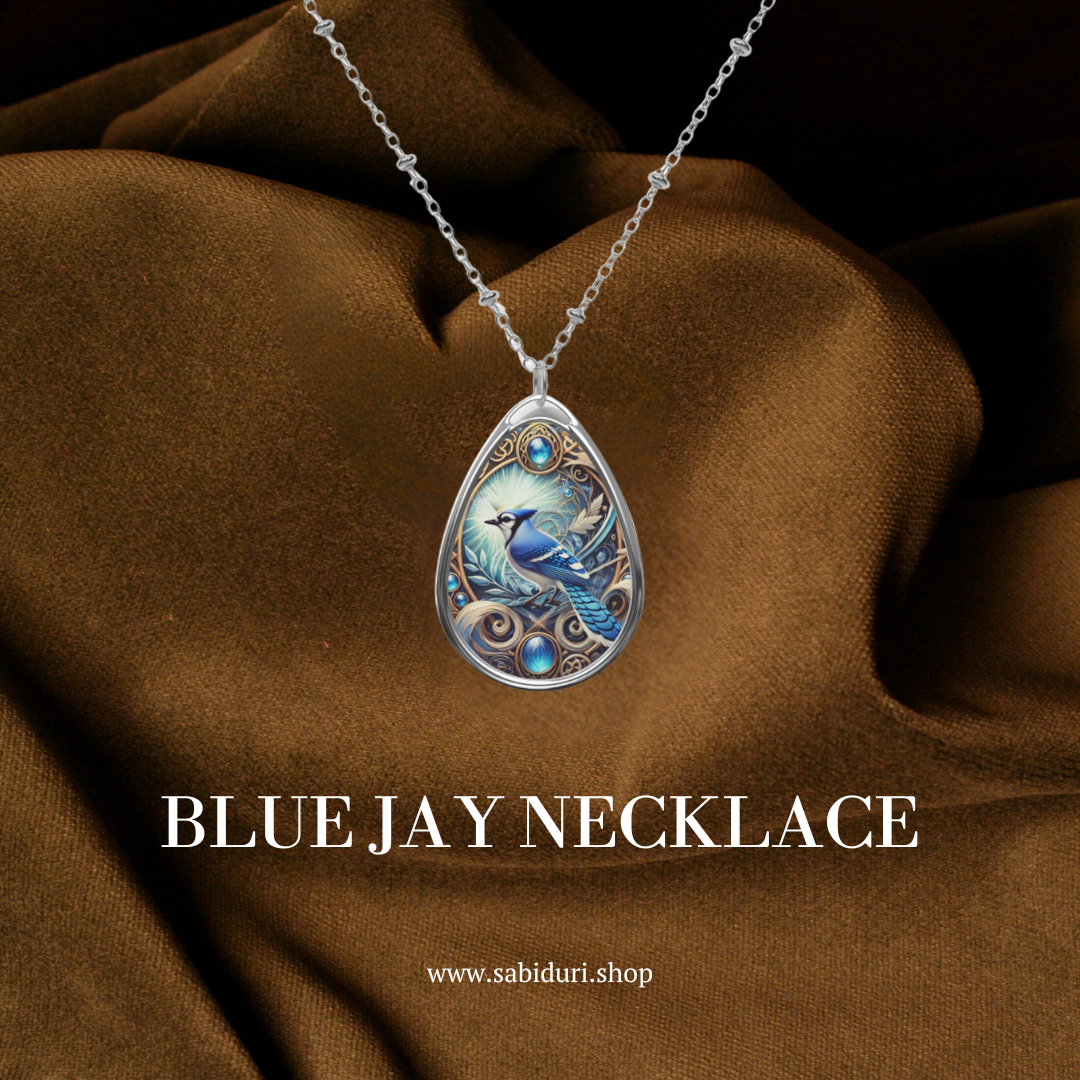 A silver teardrop pendant necklace with a detailed blue jay illustration, adorned with blue gemstones, displayed against a rich brown fabric background. The text reads "BLUE JAY NECKLACE" with the URL "www.sabiduri.shop" below.