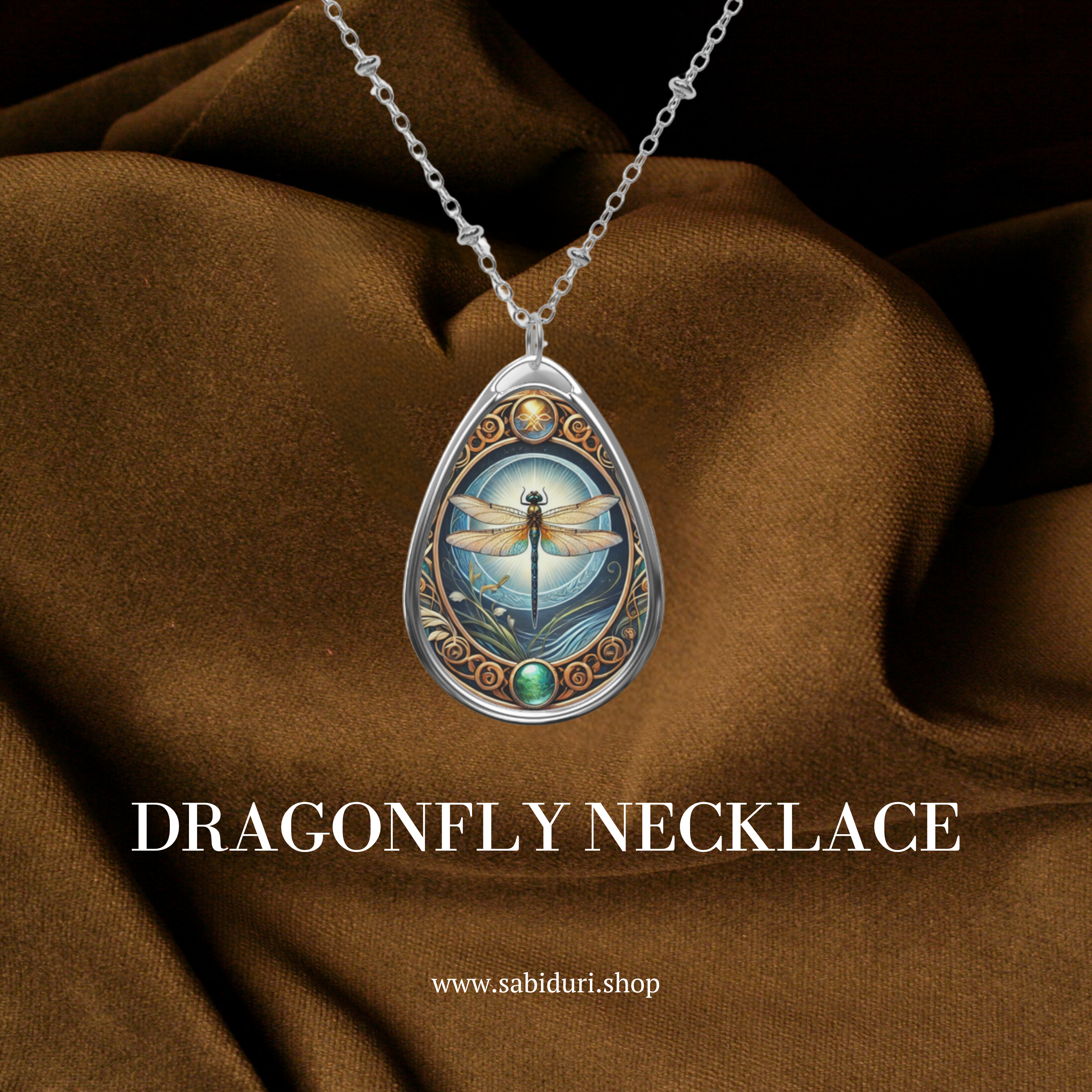 A silver dragonfly necklace with a pendant showing a dragonfly against a glowing sky and intricate golden accents, displayed against a dark brown fabric. The pendant is accented with green gemstones, and the text below reads "DRAGONFLY Necklace" with the website "www.sabiduri.shop.