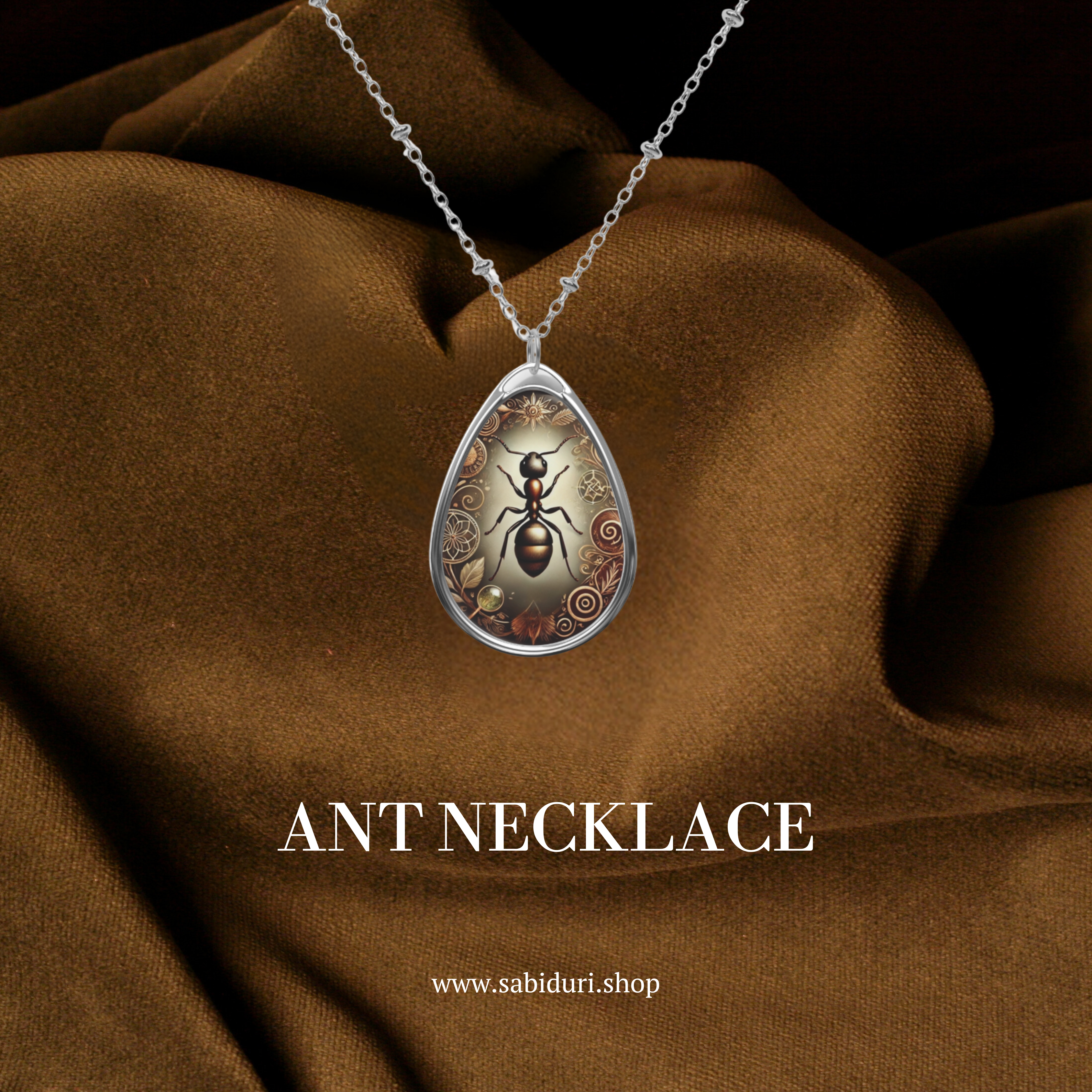 A silver ant pendant necklace with ornate engravings, featuring a highly detailed depiction of an ant against an earthy-toned background. The pendant is displayed against a luxurious brown fabric background, with the text "ANT NECKLACE" and the website "www.sabiduri.shop" below.