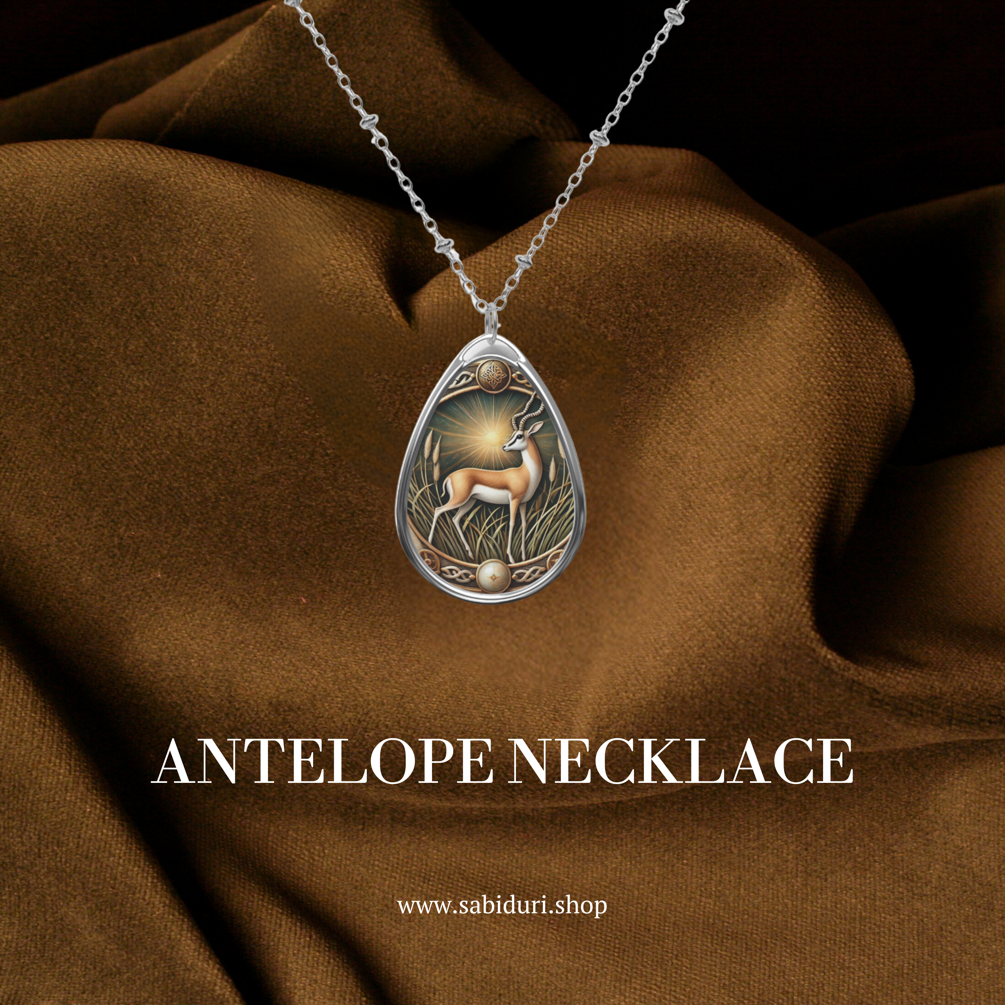 A silver antelope pendant necklace with ornate engravings, featuring a detailed depiction of an antelope in a natural setting. The pendant is displayed against a luxurious brown fabric background, with the text "ANTELOPE NECKLACE" and the website "www.sabiduri.shop" below.