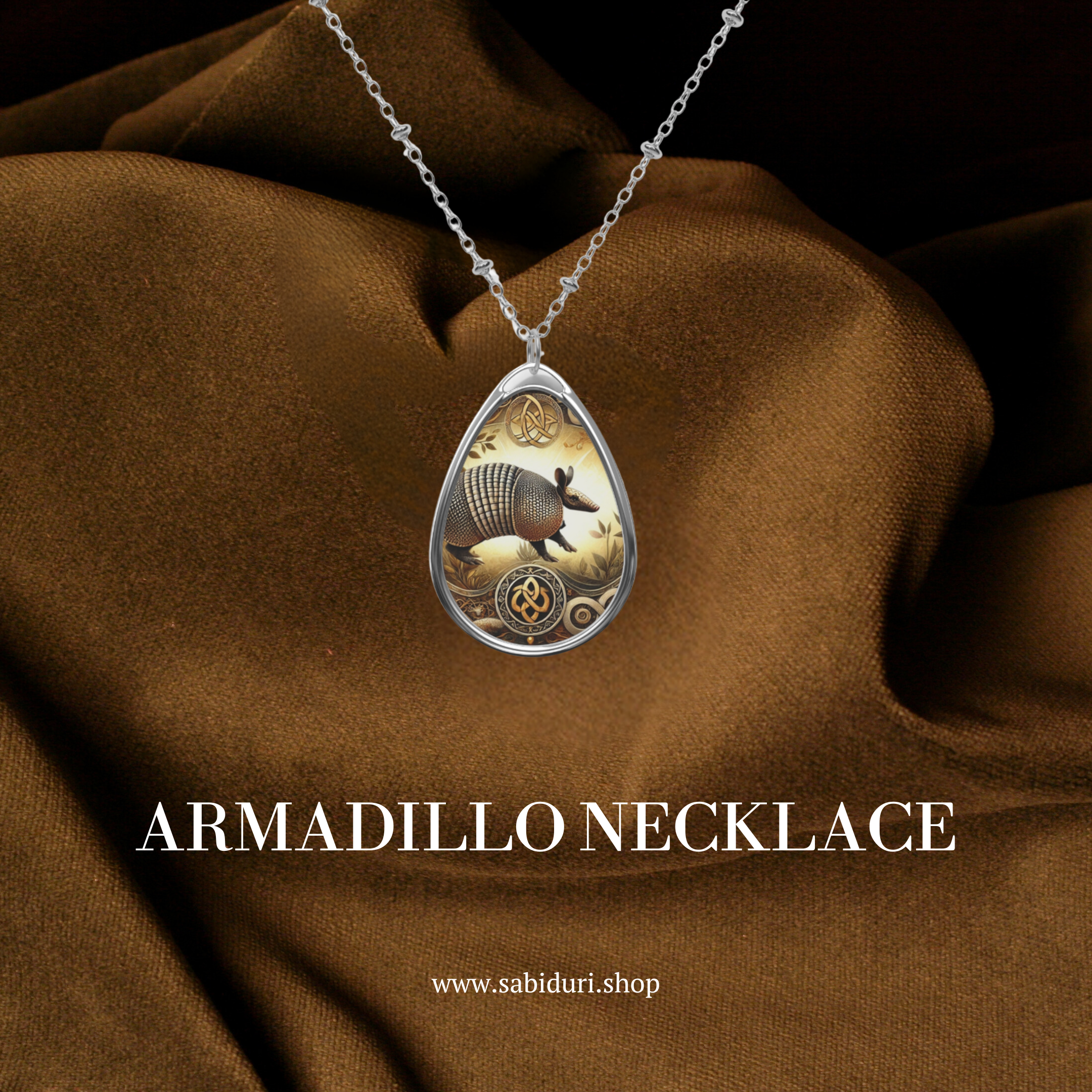 A silver chain necklace with a teardrop-shaped pendant featuring an intricately detailed armadillo illustration. The pendant is adorned with ornate engravings and displayed against a rich brown fabric backdrop, with the text "ARMADILLO NECKLACE" and the website "www.sabiduri.shop" beneath.