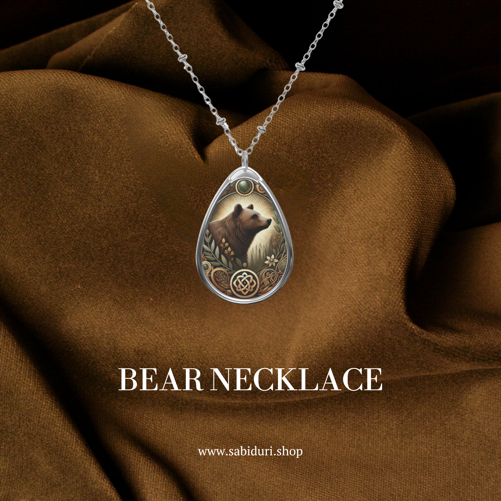 A silver chain necklace with a teardrop-shaped pendant featuring an intricate illustration of a bear surrounded by nature-inspired engravings. The pendant is set against a rich brown fabric backdrop, with the text "BEAR NECKLACE" and the website "www.sabiduri.shop" displayed below.