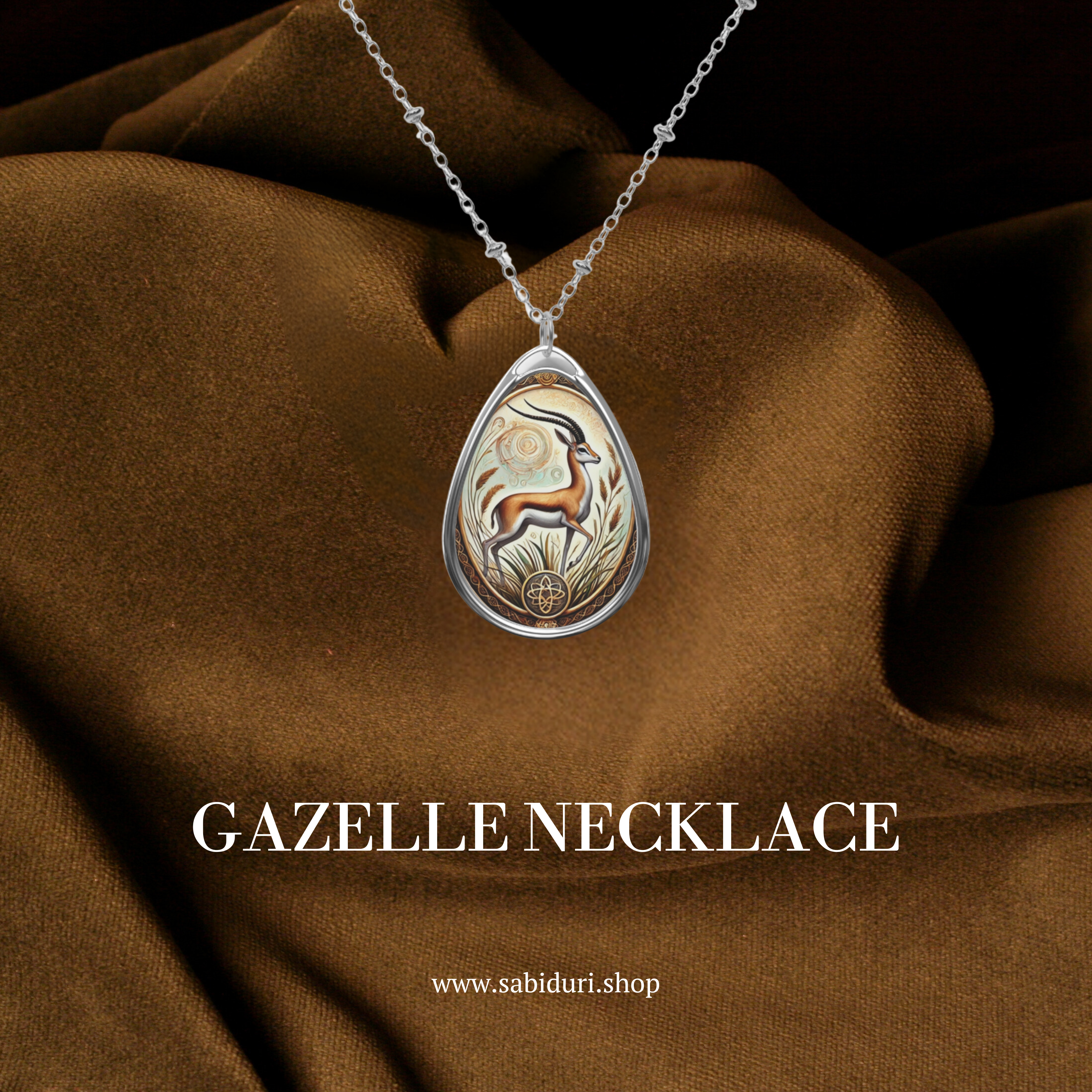 A teardrop-shaped pendant featuring an intricately designed gazelle with elegant, curved horns, standing among golden foliage. The pendant is encased in a silver frame, with delicate patterns and a circular symbol at the bottom. It is displayed on a brown fabric background with the text "GAZELLE NECKLACE" and "www.sabiduri.shop."