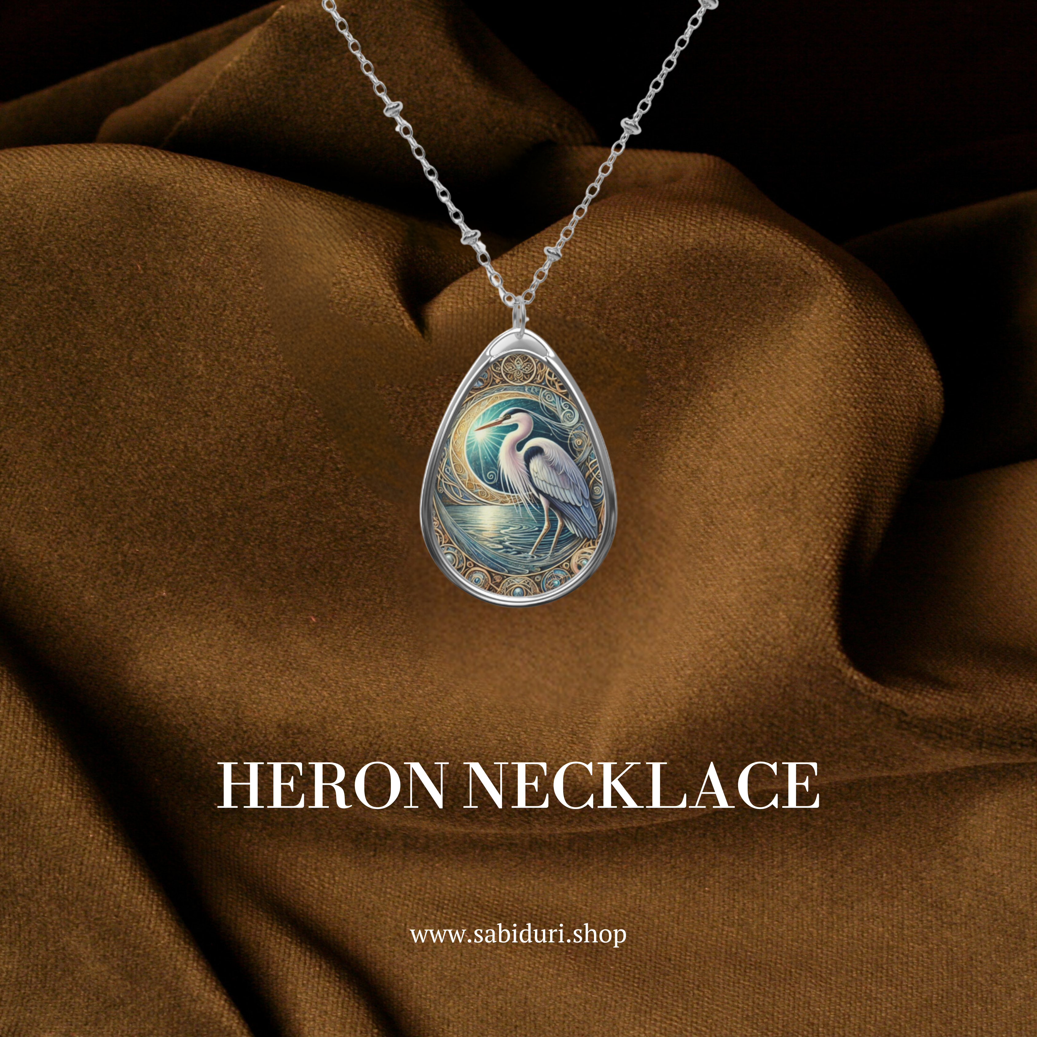 A close-up of a heron-themed necklace set against rich brown fabric. The pendant depicts a graceful heron under a glowing moon, surrounded by ornate golden patterns. The text "HERON NECKLACE" and "www.sabiduri.shop" is placed below.