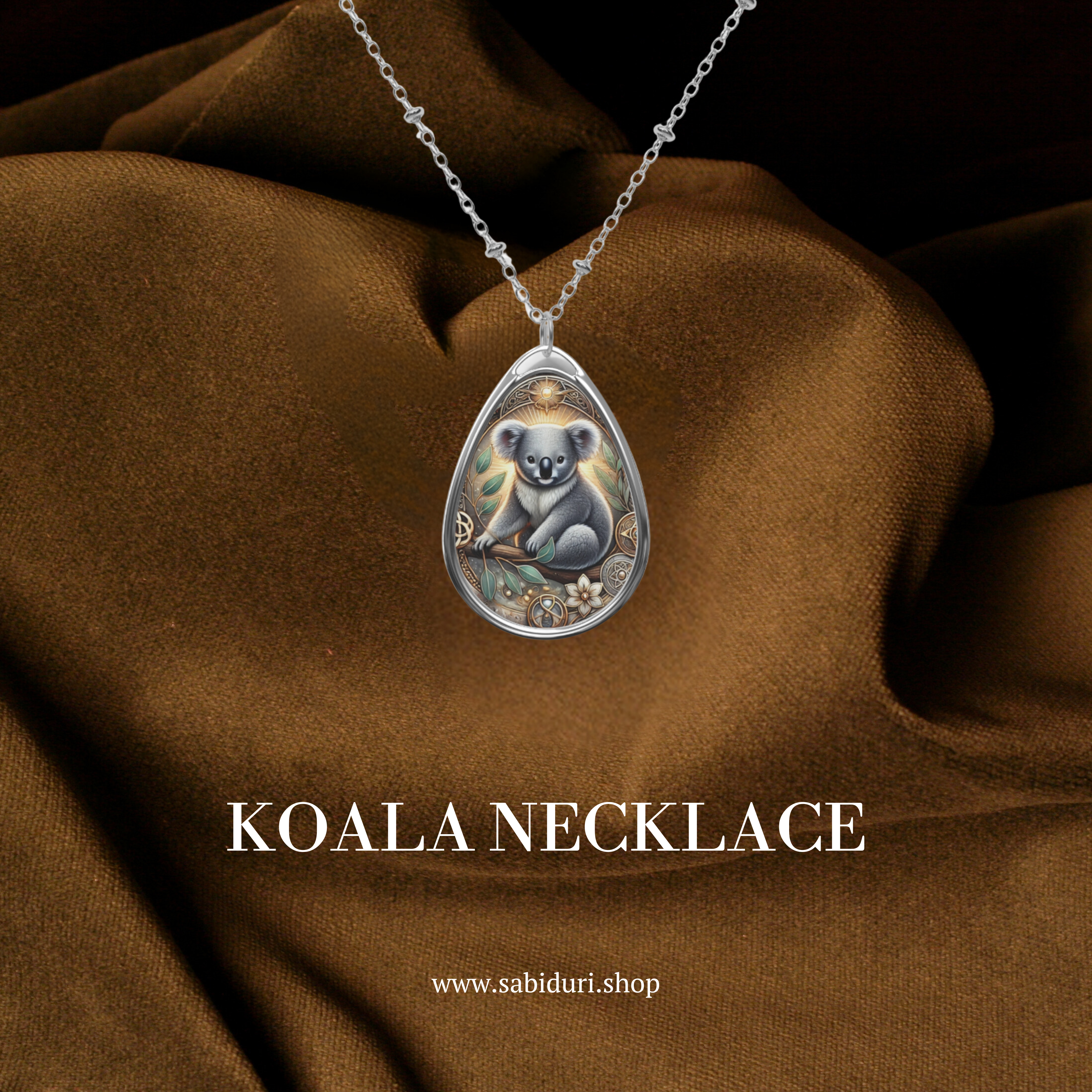 A silver teardrop pendant necklace featuring an intricately detailed koala illustration, surrounded by golden and floral accents. The necklace rests against a rich brown fabric backdrop. The text "KOALA NECKLACE" and "www.sabiduri.shop" is displayed below.