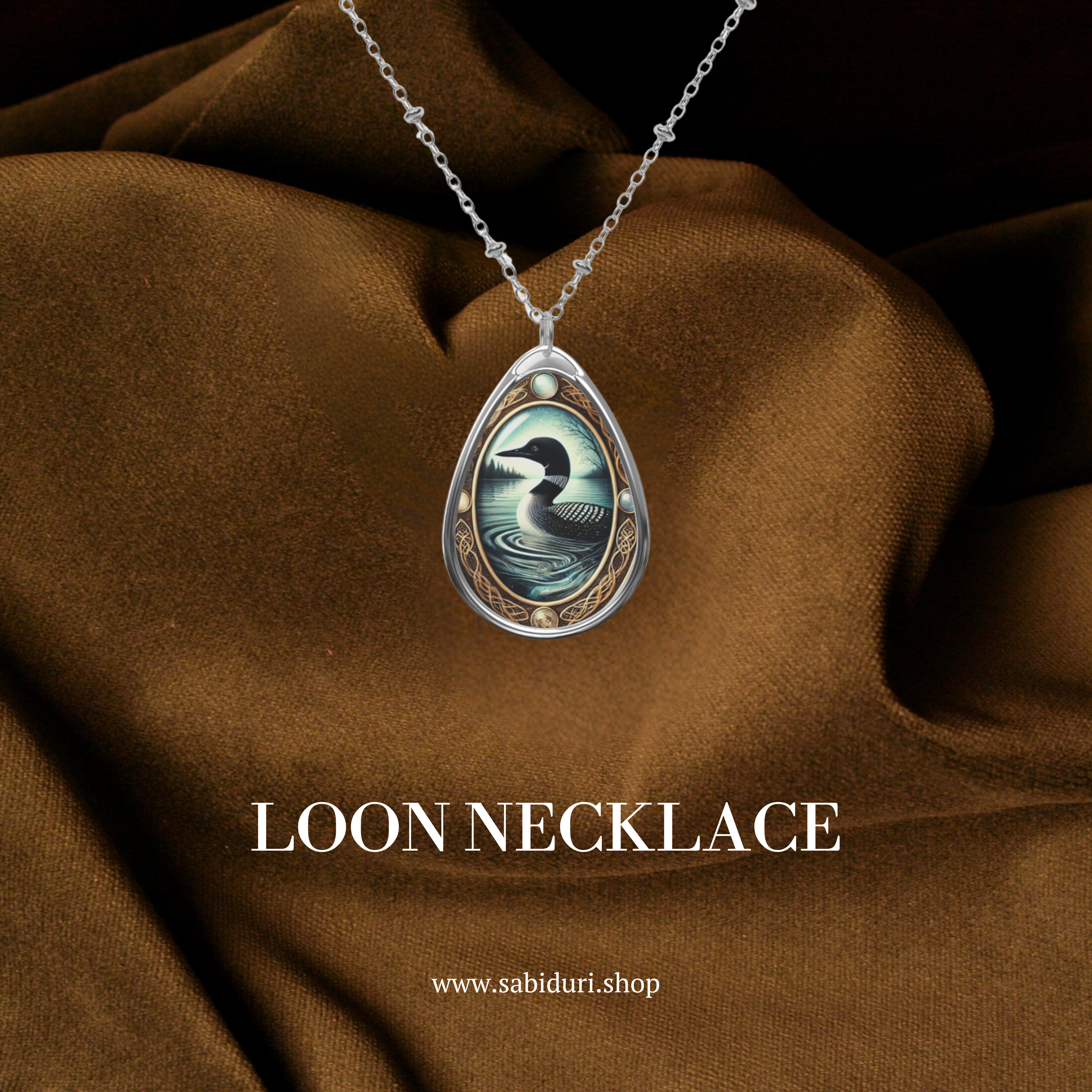 A close-up of the Loon necklace displayed against a luxurious brown fabric. The pendant's detailed artwork shows a black-and-white loon gliding through a tranquil lake, framed by ornate gold filigree. The text "LOON NECKLACE" and "www.sabiduri.shop" is elegantly placed below.