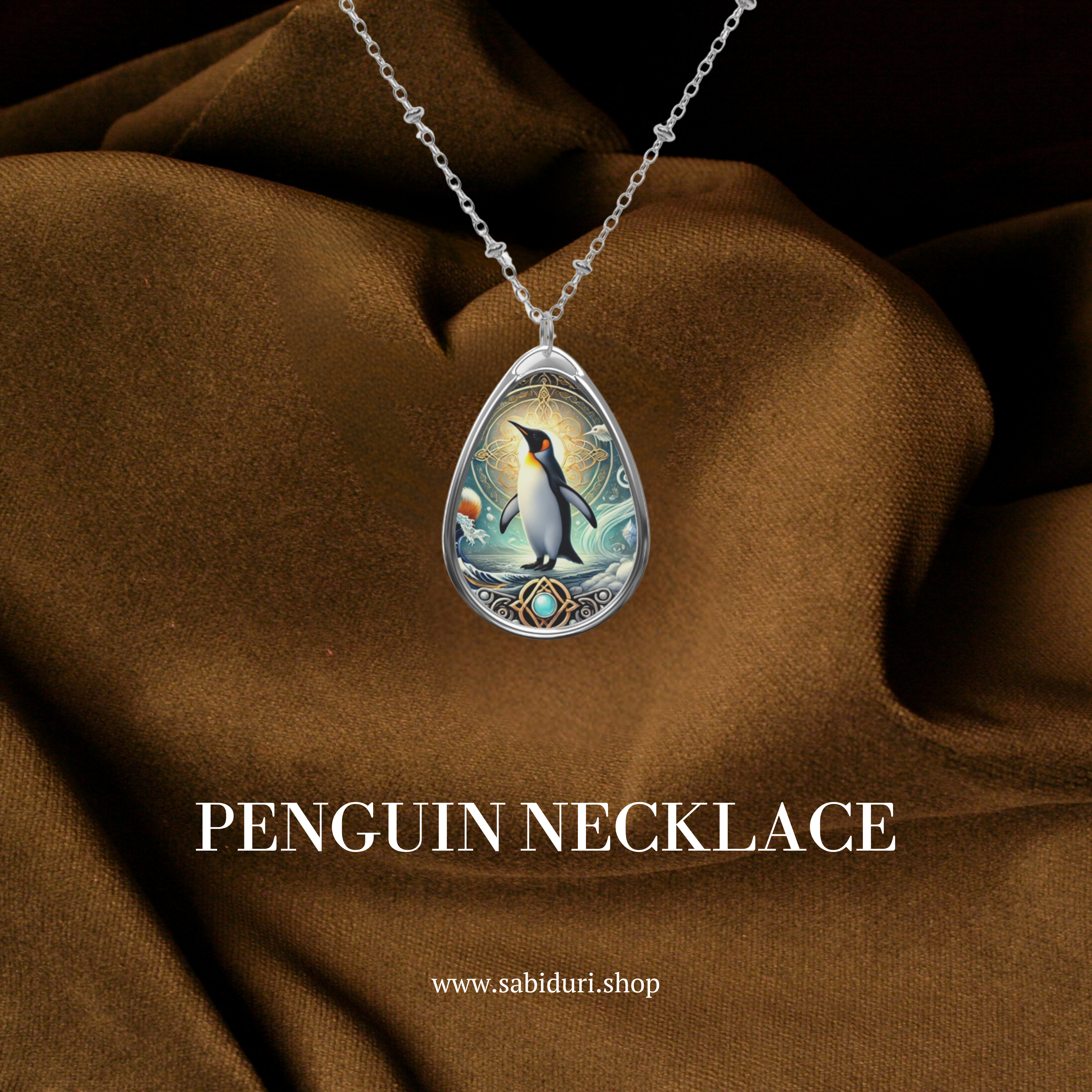 A teardrop-shaped pendant featuring an intricately designed emperor penguin standing on ice, with a golden sun and detailed engravings in the background. The necklace is displayed against a luxurious brown fabric backdrop. The text "PENGUIN NECKLACE" and "www.sabiduri.shop" is present.