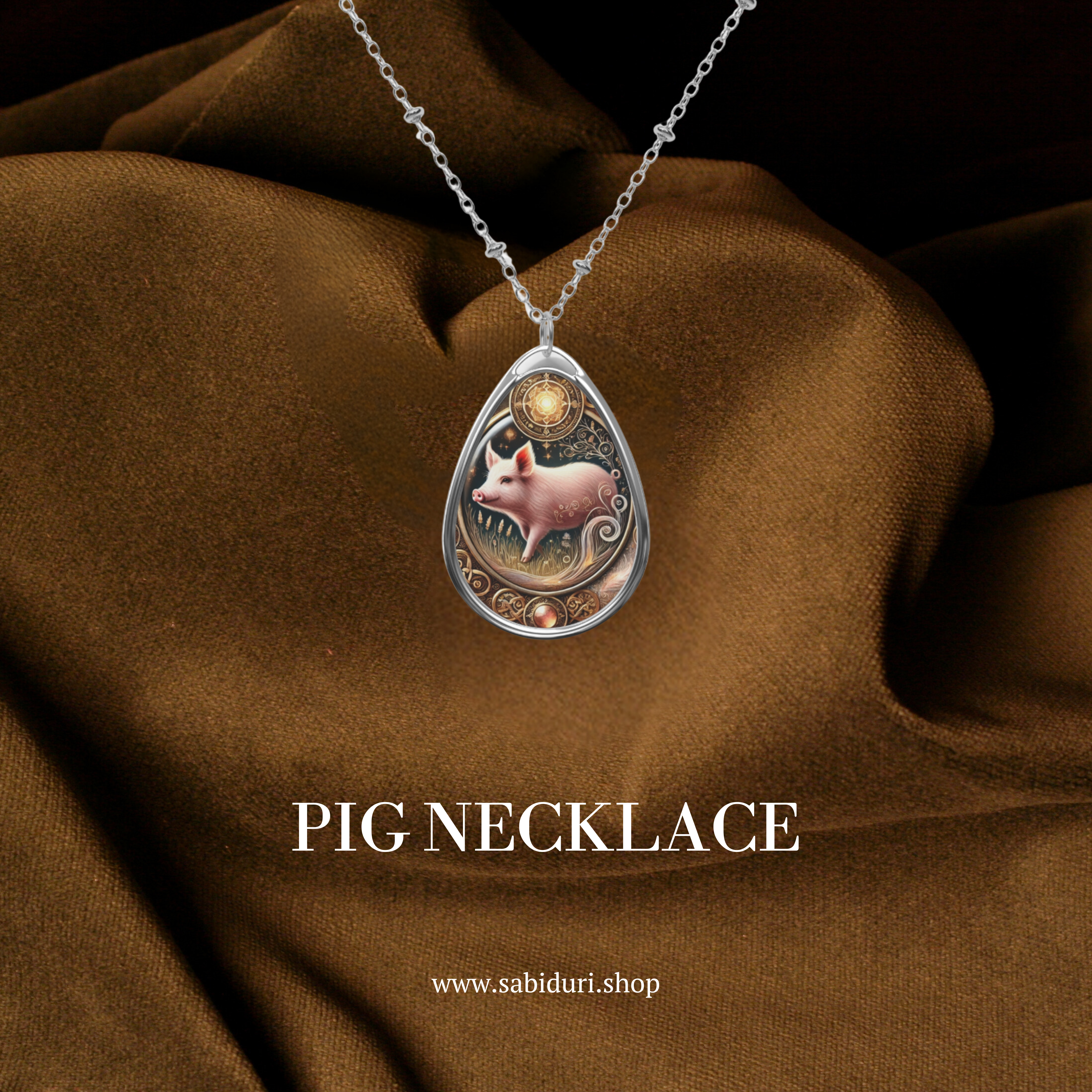 A teardrop-shaped pendant necklace featuring an intricate design of a pink pig with delicate golden swirls and floral elements. The pendant is set against a luxurious brown fabric background with the text "PIG NECKLACE" and the website "www.sabiduri.shop" displayed.