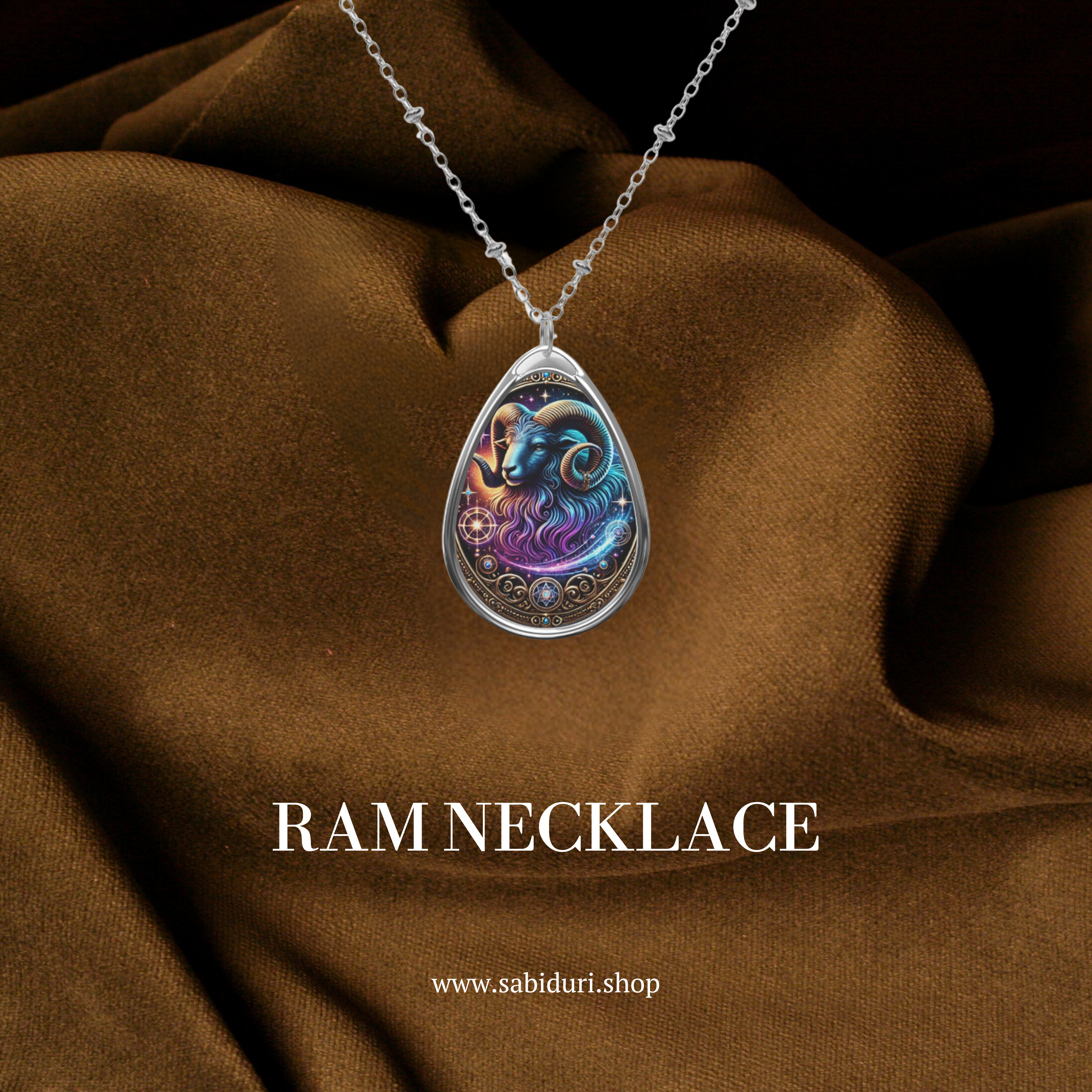 Elegant teardrop-shaped pendant featuring an intricately designed ram with swirling blue and purple hues, set against a celestial-inspired background. The pendant hangs from a delicate silver chain and is displayed on a rich brown fabric backdrop. The text 'RAM NECKLACE' and website 'www.sabiduri.shop' are included in the image.