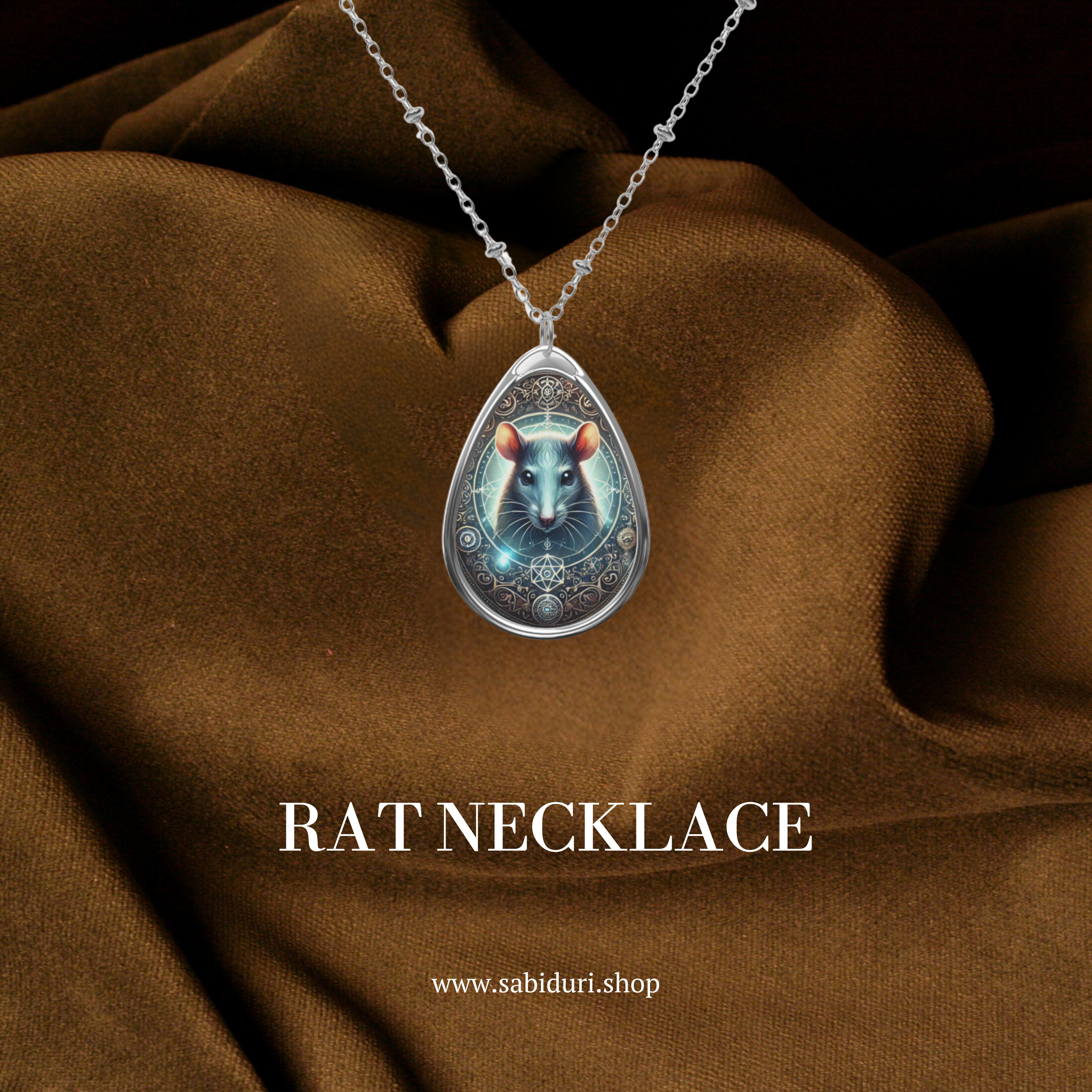 A teardrop-shaped pendant necklace featuring an intricate design of a glowing blue rat, surrounded by celestial and geometric elements. The pendant hangs on a delicate silver chain against a rich brown fabric background. Text at the bottom reads "RAT NECKLACE" and "www.sabiduri.shop."