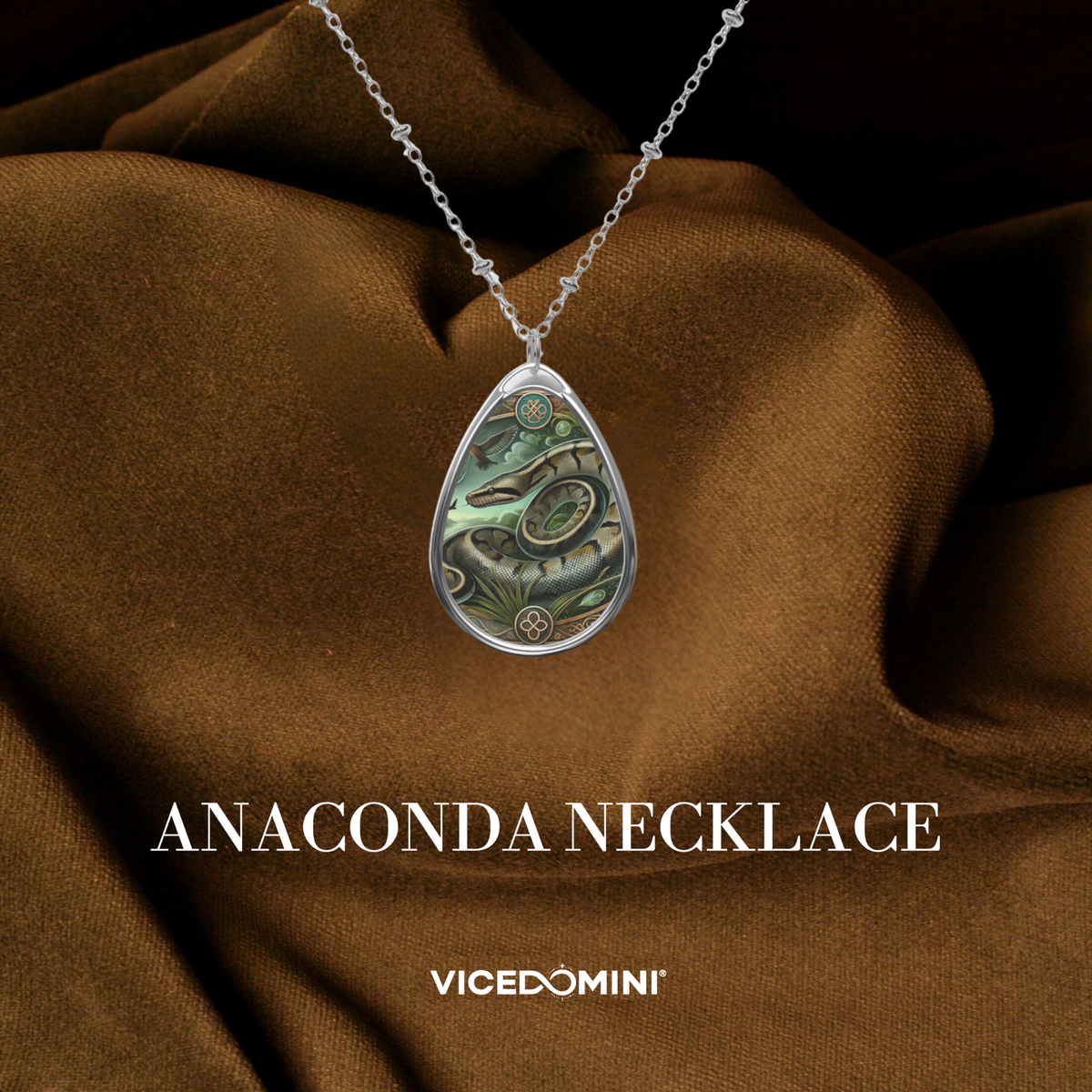 A teardrop-shaped silver pendant necklace featuring an intricate green and black snake design, displayed on draped brown fabric with the text 'ANACONDA NECKLACE' and 'VICEDOMINI' below.