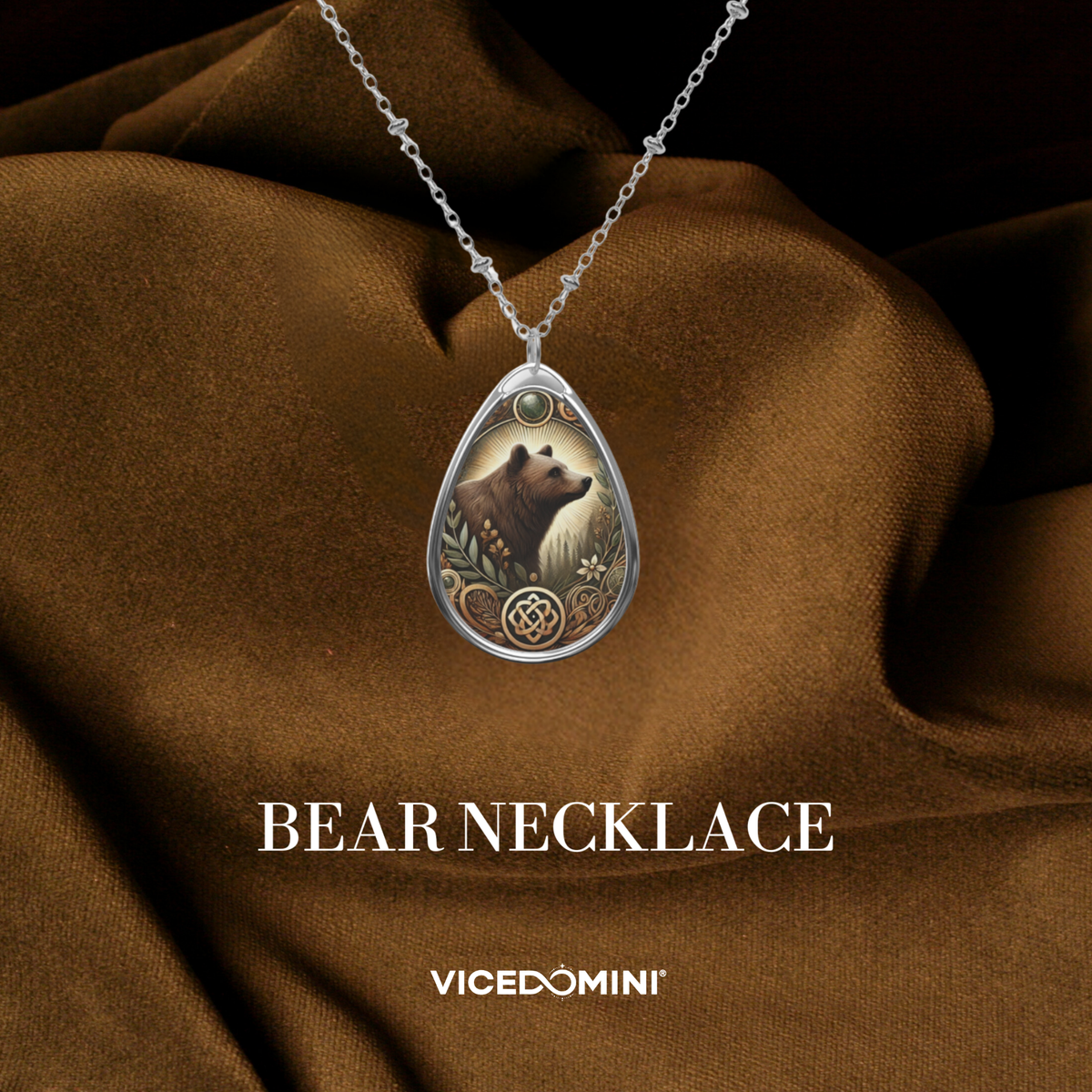 A silver teardrop pendant necklace featuring an illustrated bear surrounded by floral and geometric details, draped over brown satin fabric. The text 'BEAR NECKLACE' and the brand 'VICEDOMINI' are displayed below.