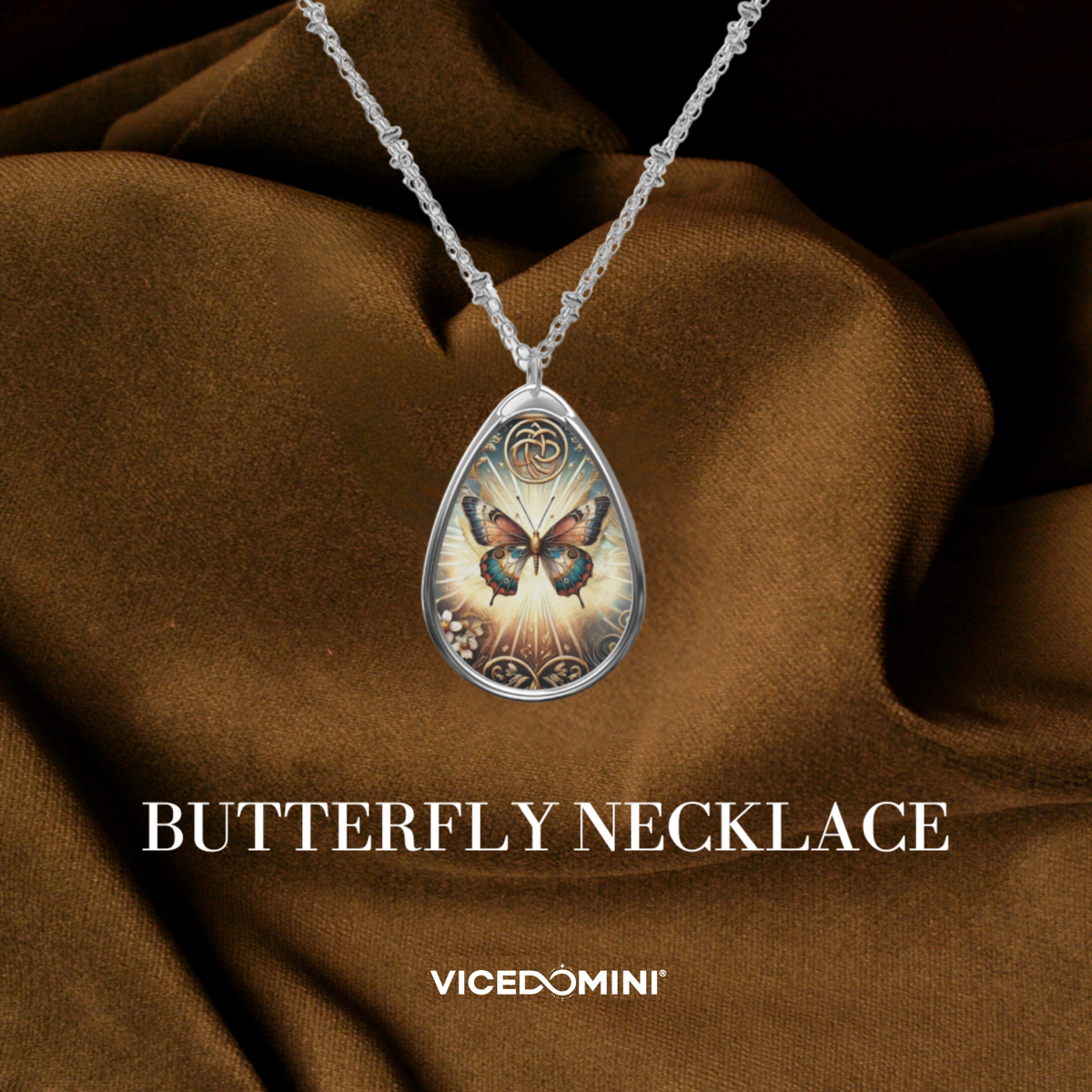 A teardrop-shaped butterfly pendant necklace on a silver chain, displayed against draped brown satin fabric. The pendant features a detailed butterfly design over a glowing light background with ornate silver borders.