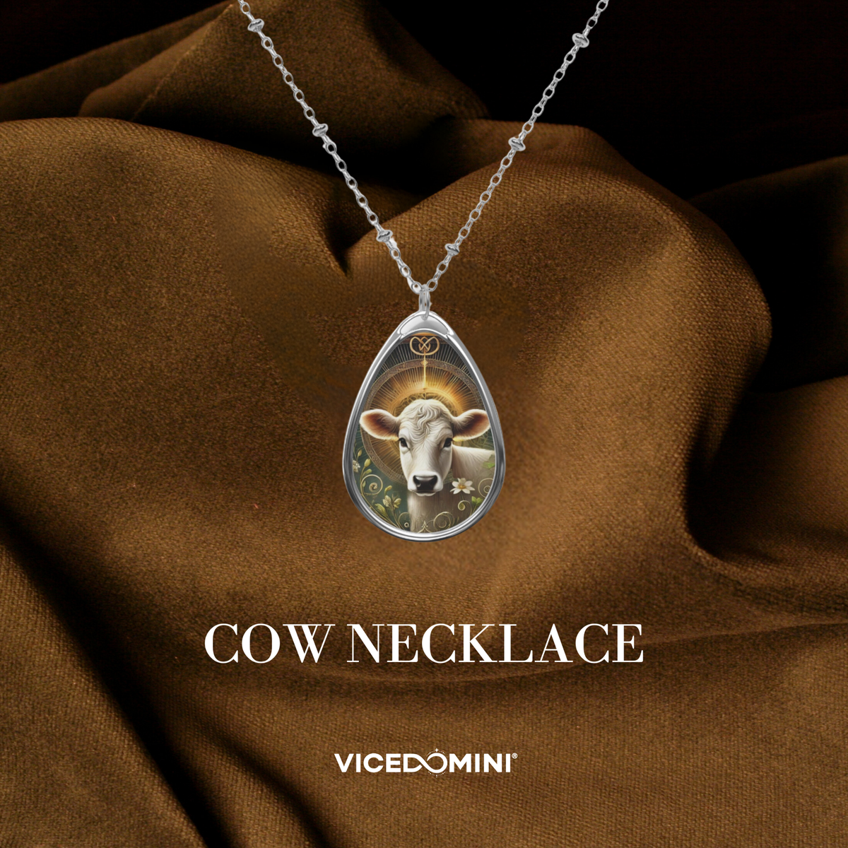 A silver teardrop pendant necklace featuring a detailed illustration of a cow with a halo, draped over brown satin fabric. The text 'COW NECKLACE' and the brand 'VICEDOMINI' are displayed below.