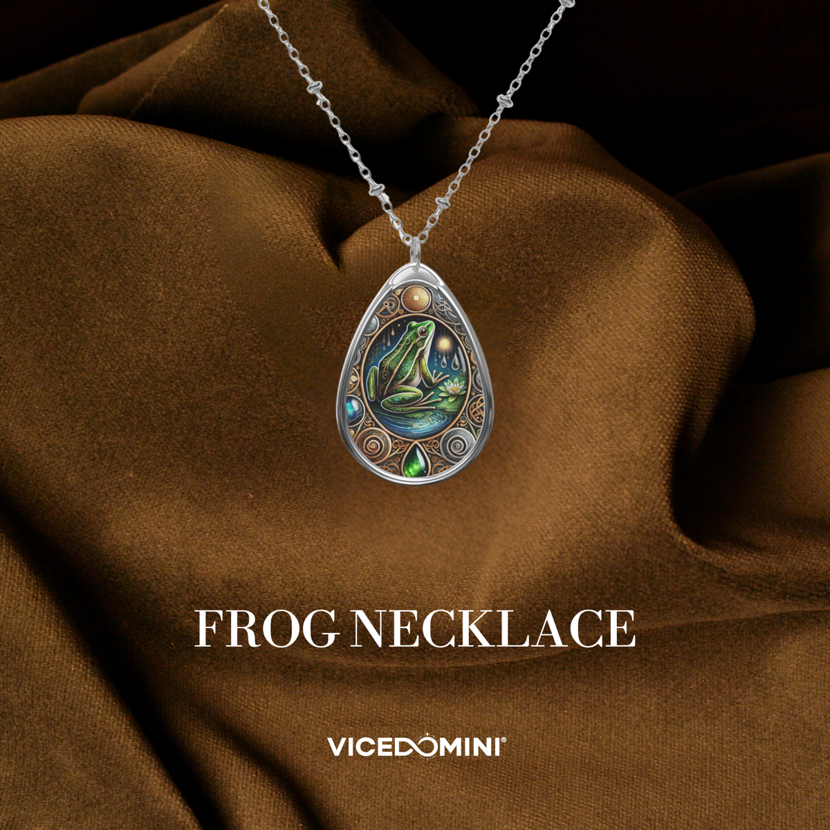 A teardrop-shaped frog pendant necklace with a silver-toned chain, displayed against draped brown fabric. The pendant features a detailed frog illustration with green, blue, and gold accents, surrounded by decorative silver filigree.