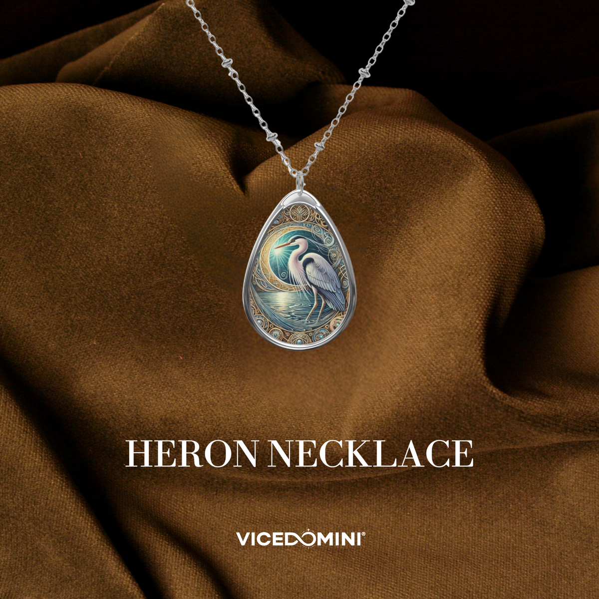 A teardrop-shaped silver pendant necklace featuring an intricate illustration of a heron in blue, white, and gold tones, displayed on draped brown fabric with the text 'HERON NECKLACE' and 'VICEDOMINI' below.