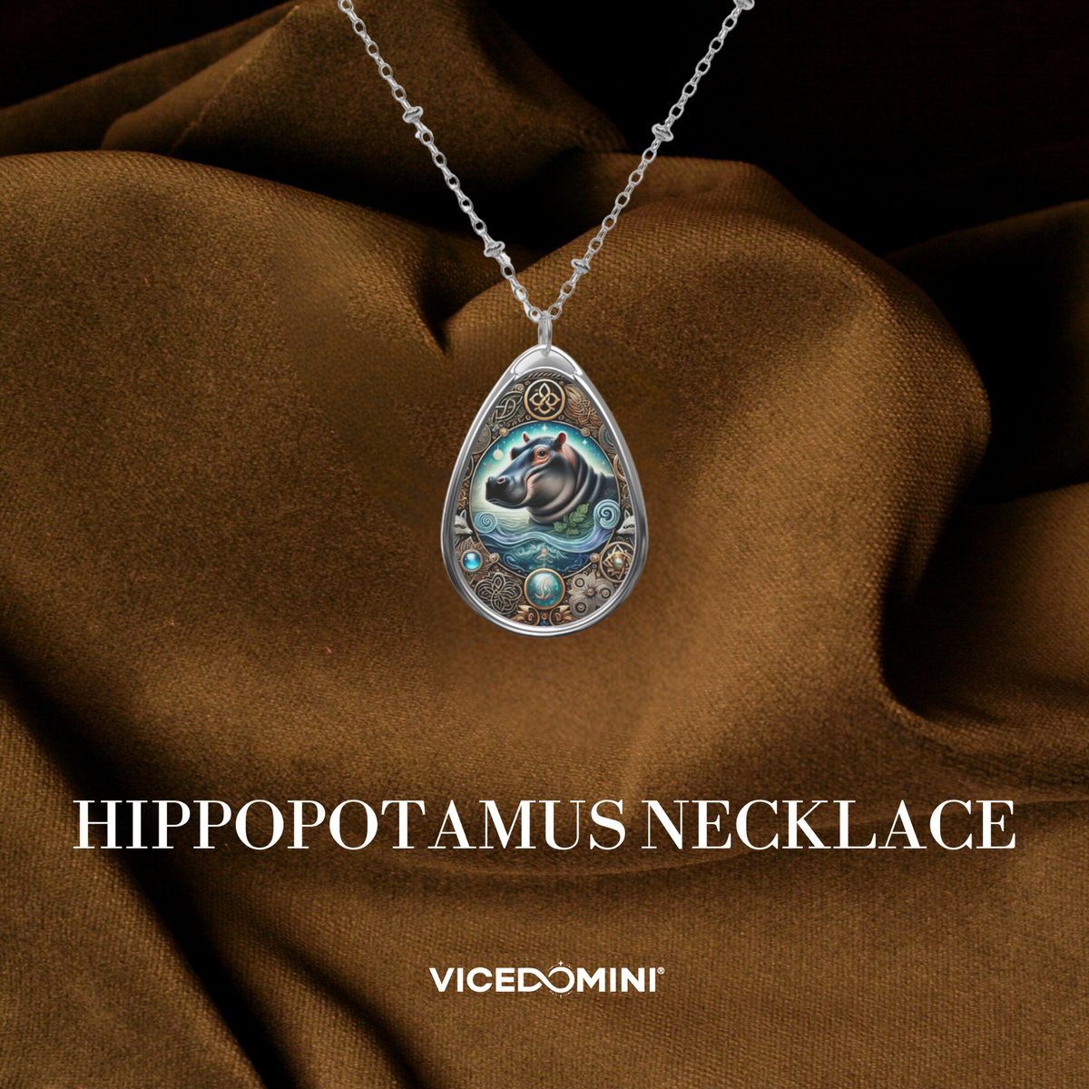 A teardrop-shaped silver pendant necklace featuring an illustrated hippopotamus surrounded by ornate circular details, hanging from a silver chain against draped brown fabric. The text 'HIPPOPOPOTAMUS NECKLACE' and the brand 'VICEDOMINI' are displayed below.