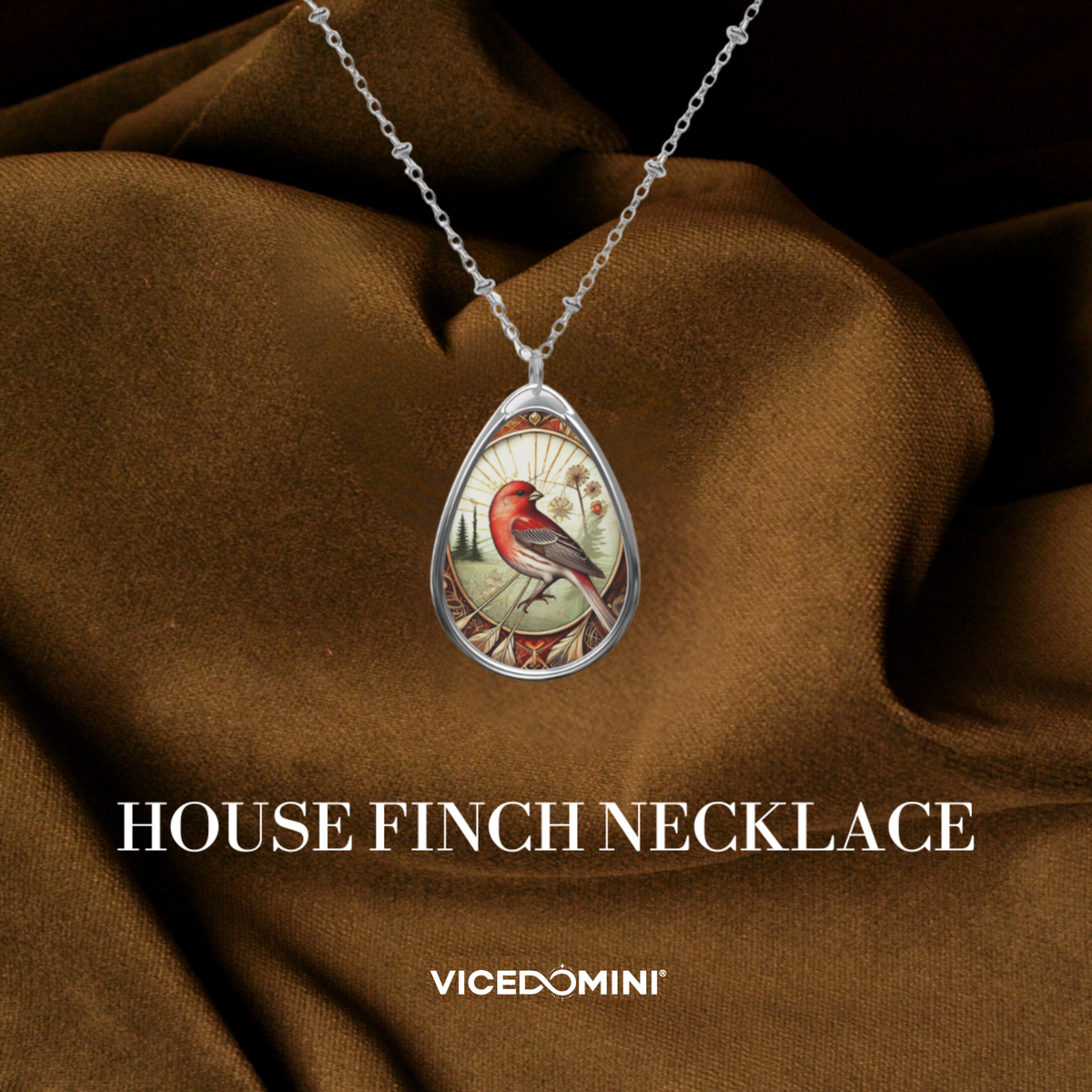 A silver teardrop pendant necklace featuring a detailed illustration of a red house finch, draped over brown satin fabric. The text 'HOUSE FINCH NECKLACE' and the brand 'VICEDOMINI' are displayed below.