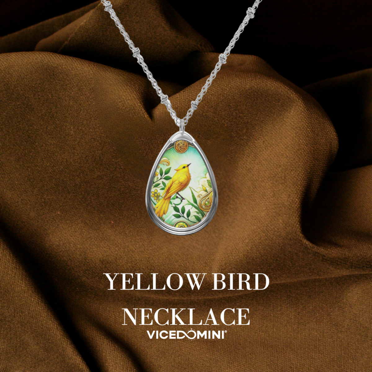 A teardrop-shaped pendant necklace featuring a yellow bird illustration on a light green background, suspended from a silver chain over draped brown fabric. The text 'YELLOW BIRD NECKLACE' and 'VICEDOMINI' are displayed below.