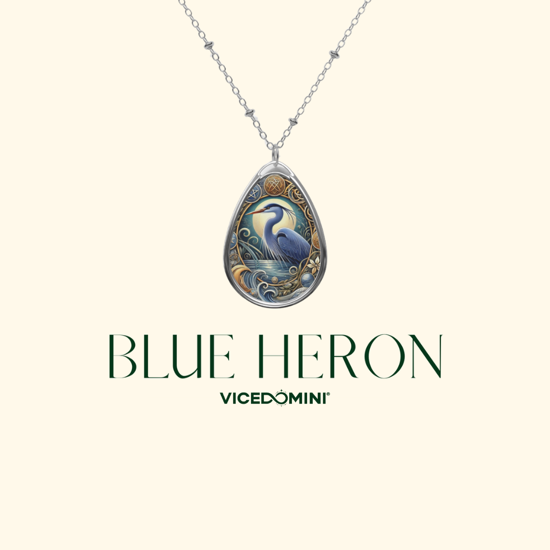 A teardrop-shaped pendant necklace featuring an illustrated blue heron with ornate gold and blue detailing, suspended from a silver-toned chain against a pale cream background. The text 'BLUE HERON' and 'VICEDOMINI' appears below the pendant.