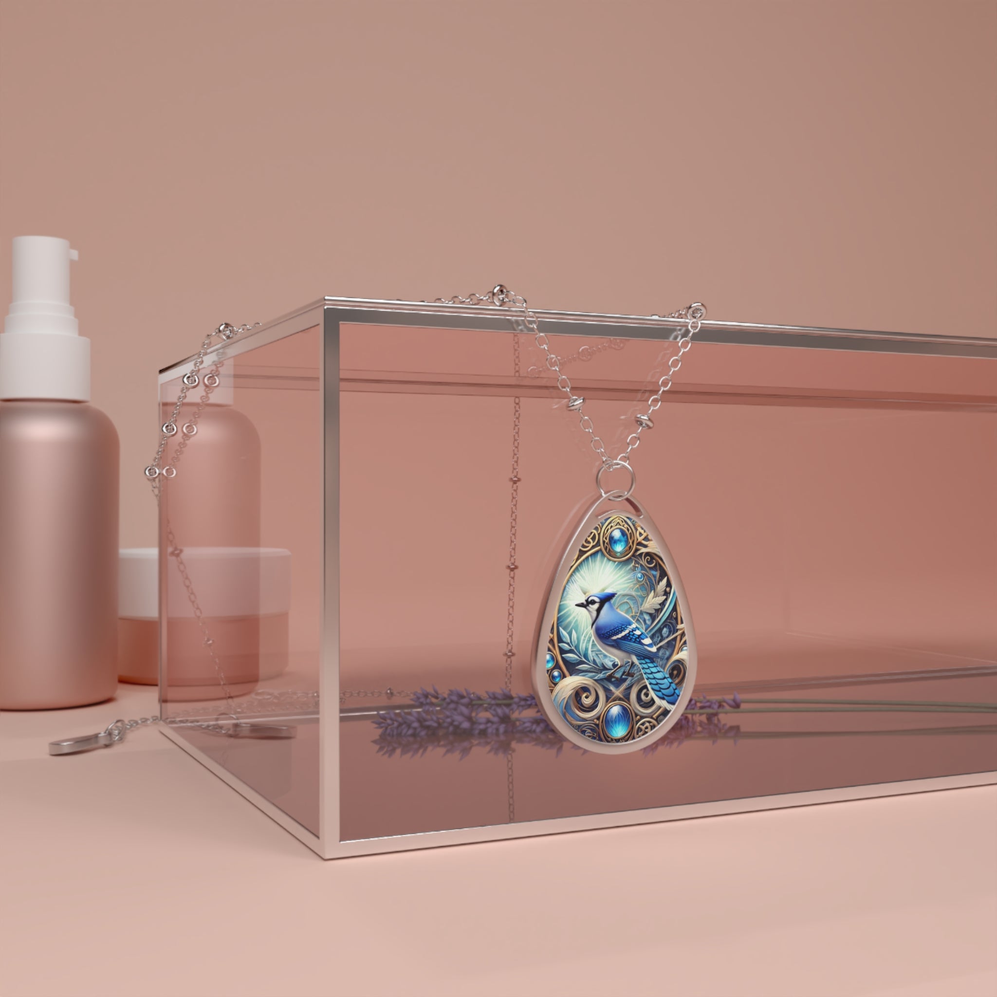 A silver necklace featuring an ornate teardrop pendant with a detailed illustration of a blue jay, accented with blue gemstones. The necklace is draped over a clear, rectangular display box with rose-gold beauty products in the background.