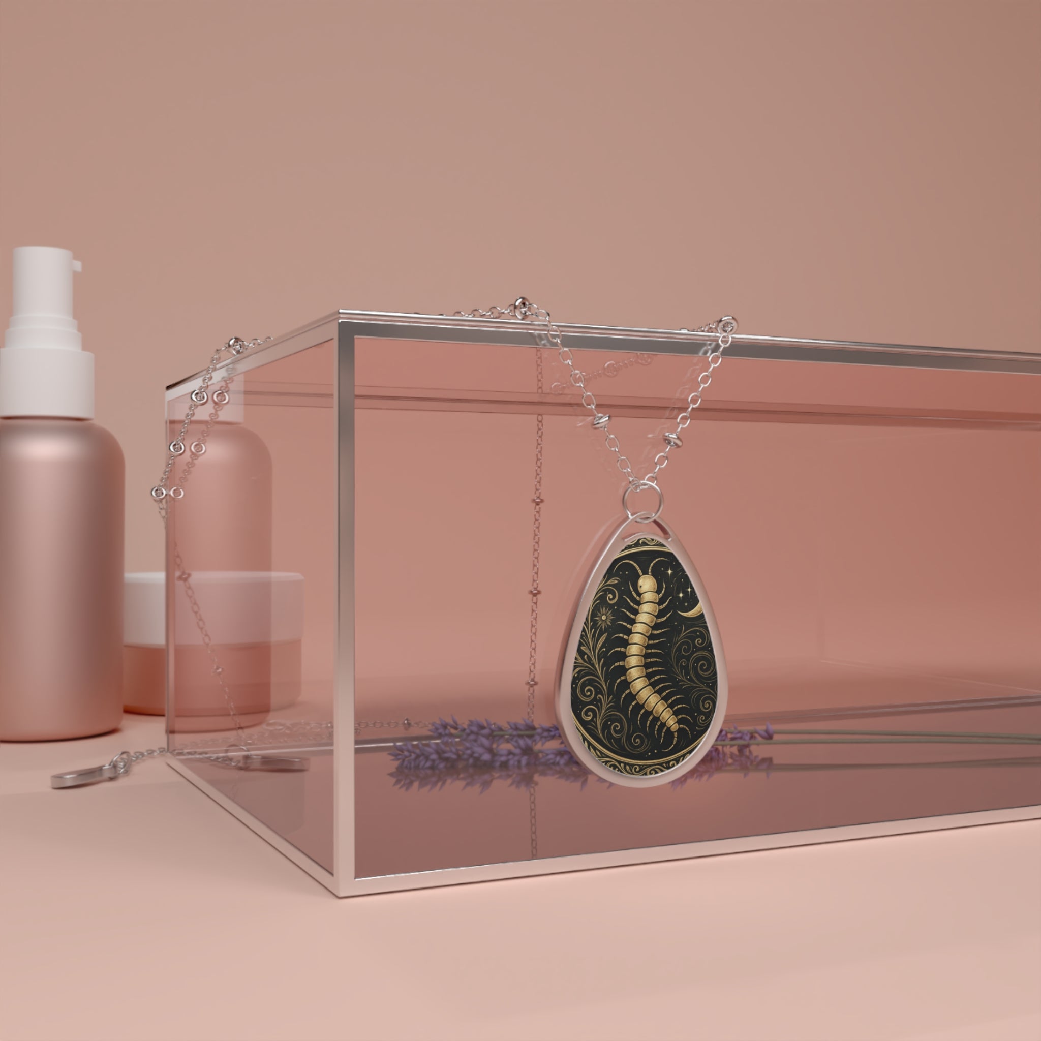 Teardrop-shaped necklace with a gold centipede design set against a swirling black background, draped over a pink glass box beside lavender and skincare items.