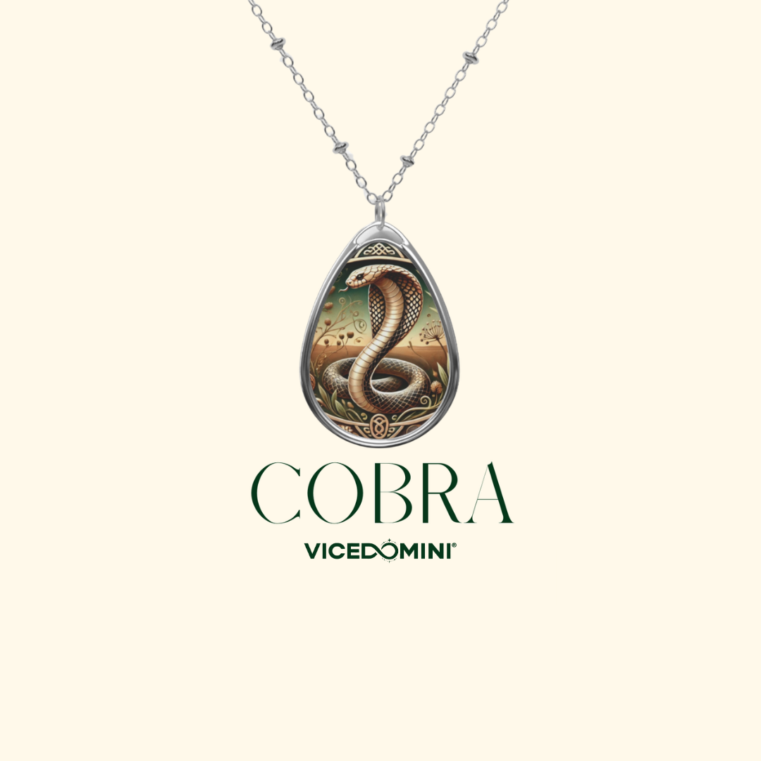 A silver teardrop pendant necklace featuring an illustrated cobra snake design, suspended from a silver chain against a light cream background. Below the necklace, the brand name 'COBRA VICEDOMINI' is printed in dark green.
