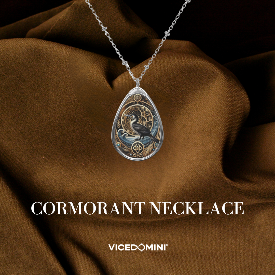 A teardrop-shaped silver pendant necklace featuring a cormorant illustration with blue-gray and gold details, displayed on draped brown fabric. The text 'CORMORANT NECKLACE' and the brand 'VICEDOMINI' are visible at the bottom.