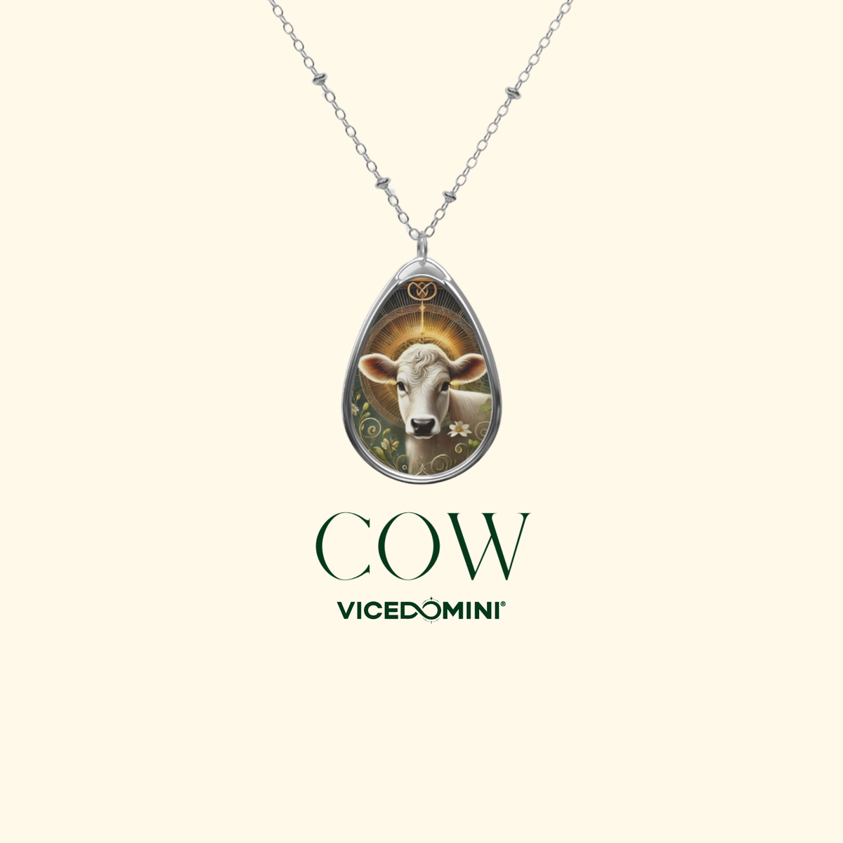 A silver teardrop pendant necklace featuring a detailed illustration of a white cow with a golden halo, shown on a light cream background with the text 'COW VICEDOMINI' below.