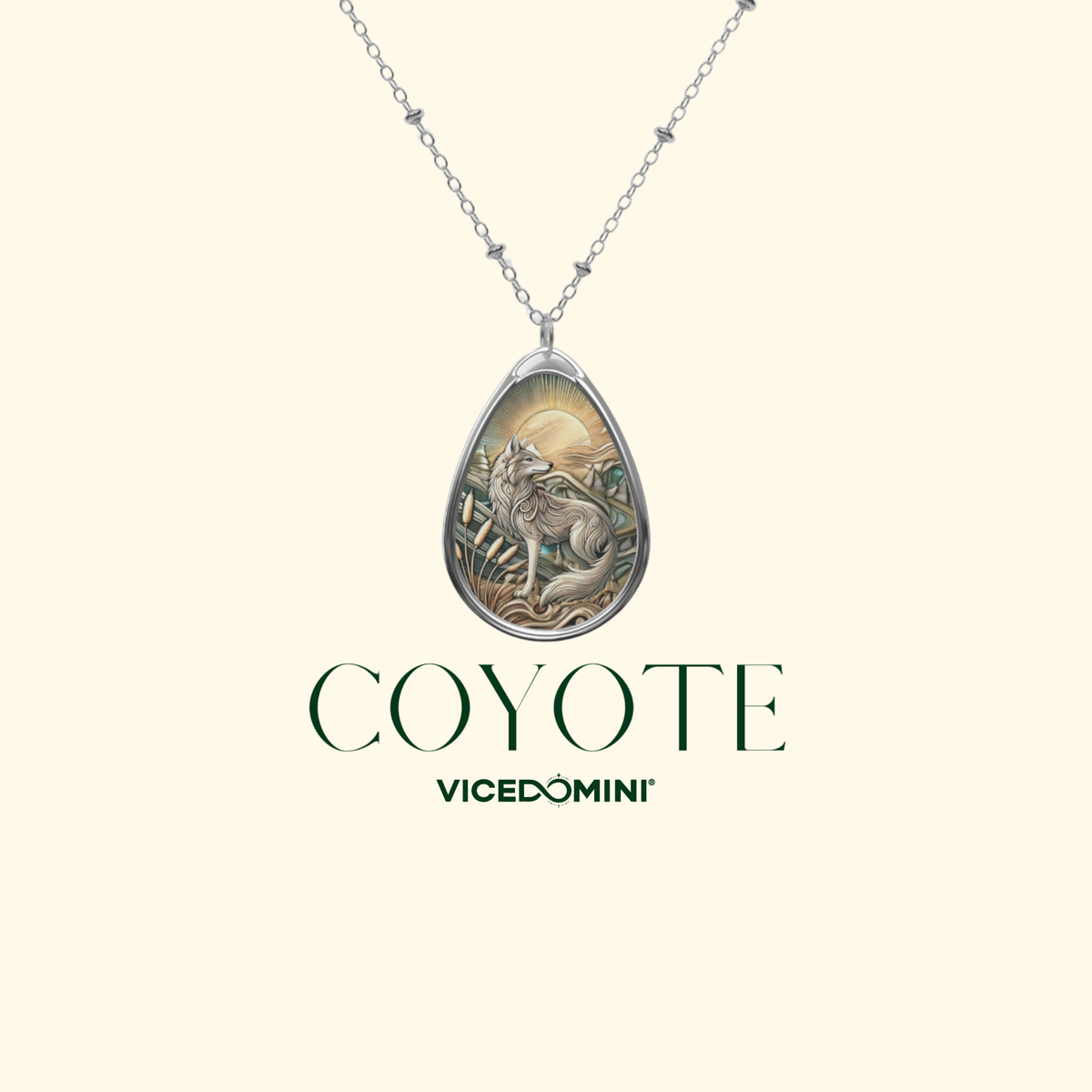 A silver teardrop pendant necklace featuring an engraved coyote illustration with a warm-toned sunlit background, shown on a chain against a pale cream backdrop. The words 'COYOTE' and 'VICEDOMINI' appear below the pendant.