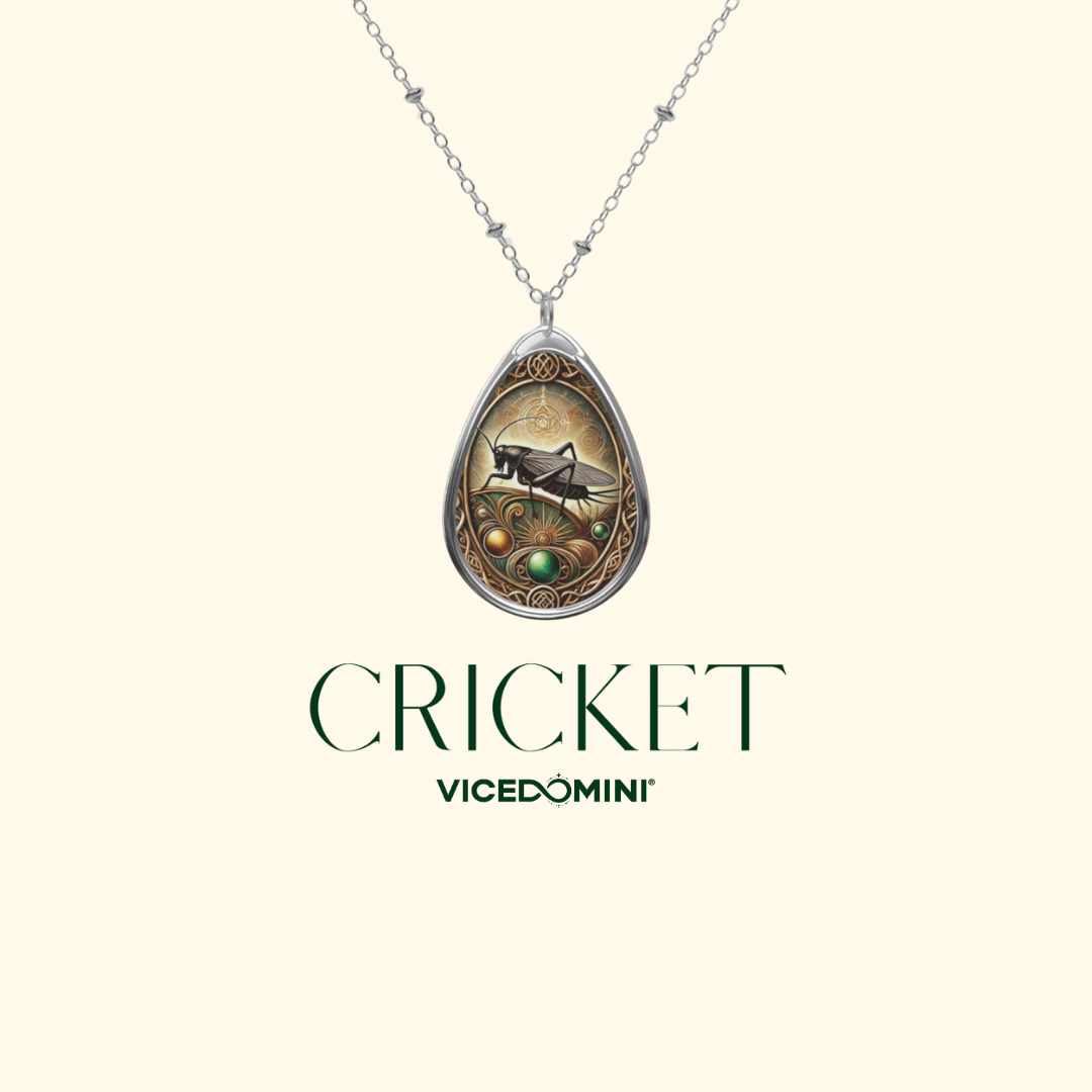 A silver teardrop pendant necklace featuring an illustrated cricket in a decorative frame, shown on a light cream background with the text 'CRICKET VICEDOMINI' below.