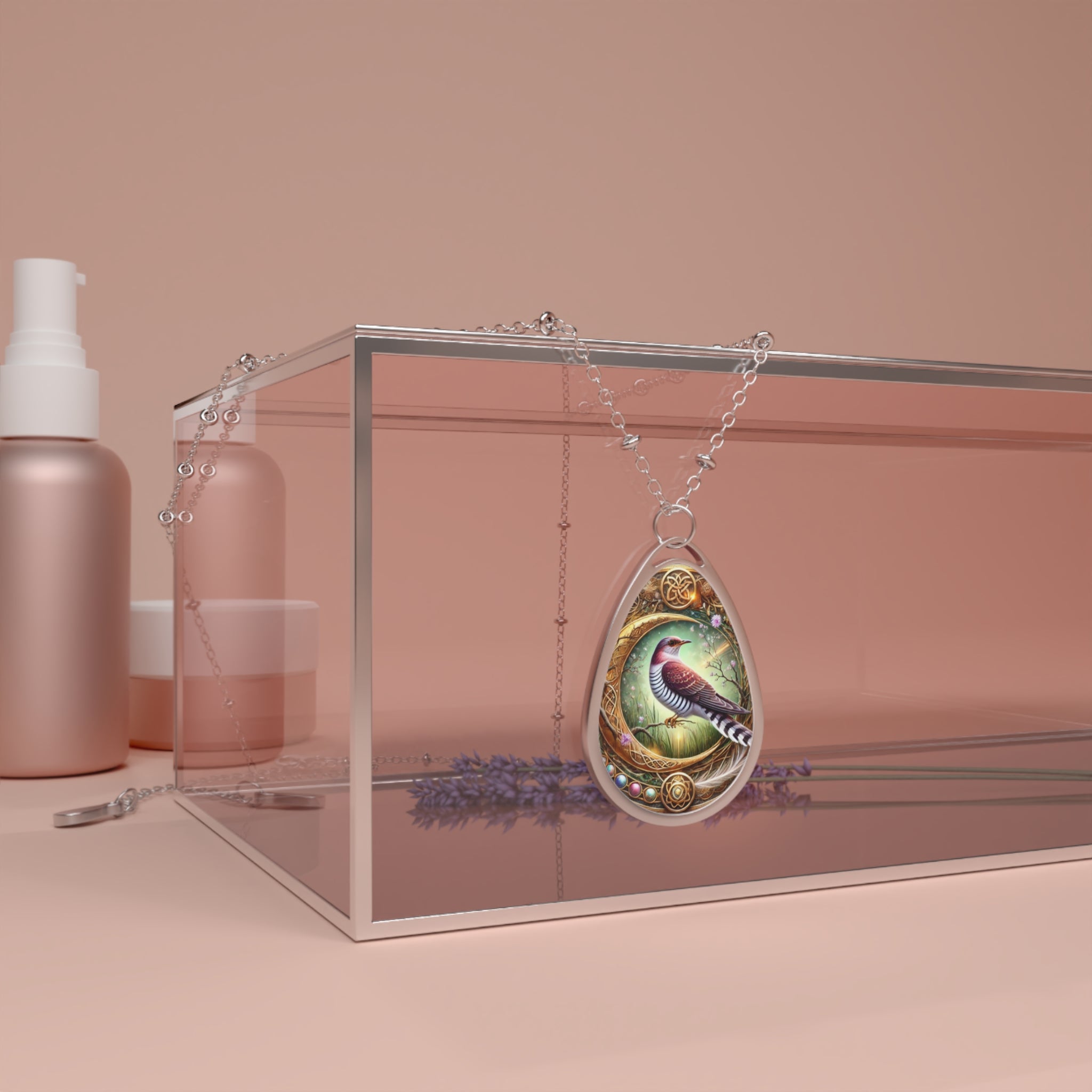 A close-up of a silver teardrop pendant necklace featuring an intricate design of a cuckoo bird perched in a golden, nature-inspired frame, displayed on a pink glass box with lavender sprigs below.