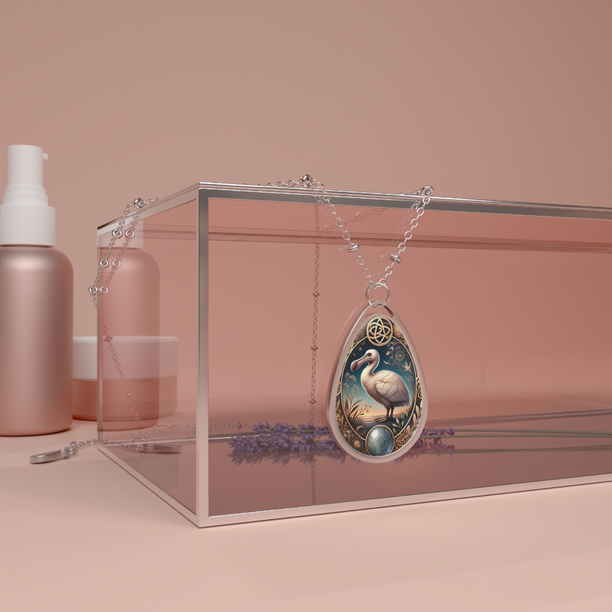 A teardrop-shaped pendant featuring an intricate illustration of a dodo bird, surrounded by celestial and botanical details. The silver necklace drapes over a transparent box with a warm-toned background, with lavender sprigs subtly reflected inside.