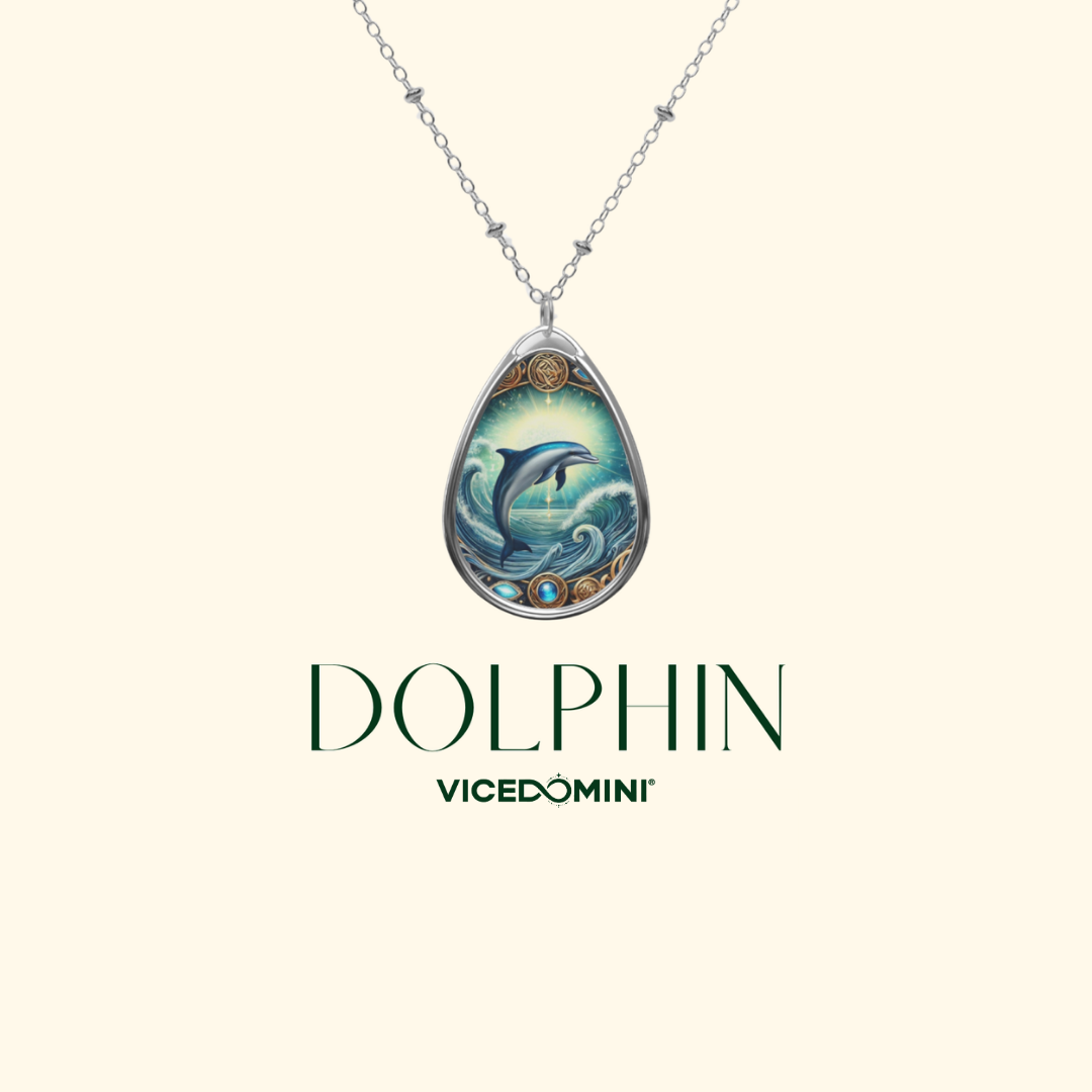 A teardrop-shaped pendant necklace featuring an illustrated dolphin in blue ocean waves, suspended from a silver-toned chain against a light cream background. The word 'DOLPHIN' appears below the pendant, with 'VICEDOMINI' underneath in dark green text.