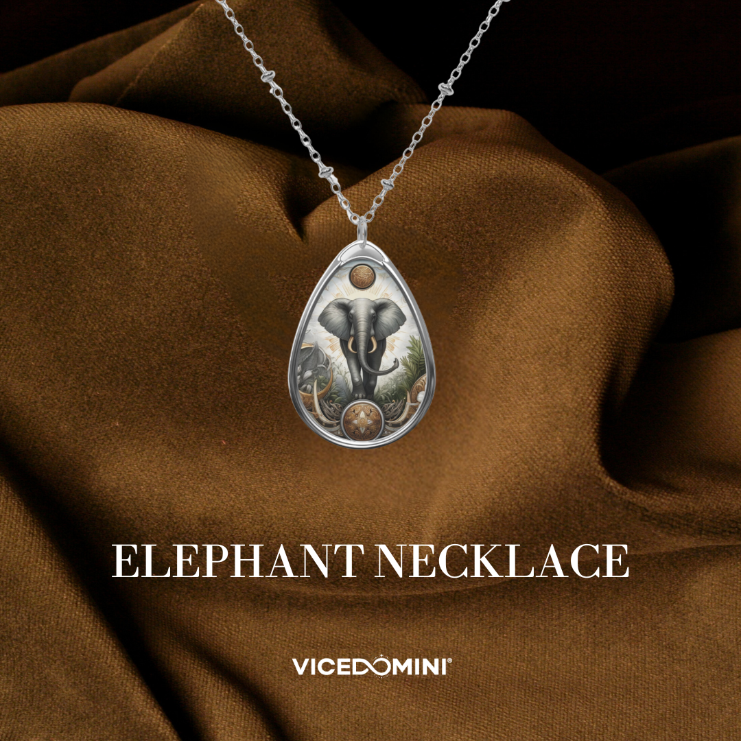 A teardrop-shaped silver pendant necklace featuring an elephant illustration, draped over brown satin fabric. The text 'ELEPHANT NECKLACE' and the brand 'VICEDOMINI' are displayed below.