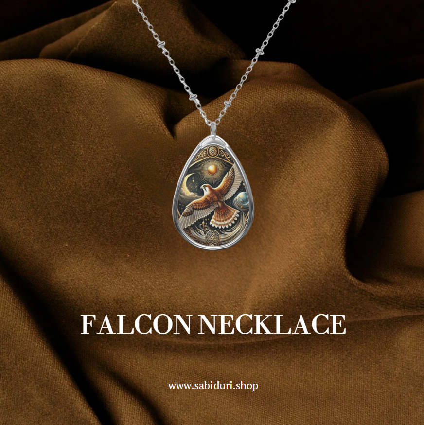 A teardrop-shaped pendant necklace featuring an intricately detailed falcon soaring with outstretched wings against a celestial background with a glowing sun, moon, and planets. The pendant is framed with golden ornamental designs and small gemstones, hanging on a delicate silver chain over a rich brown fabric. Below, the text reads "FALCON NECKLACE" and "www.sabiduri.shop."