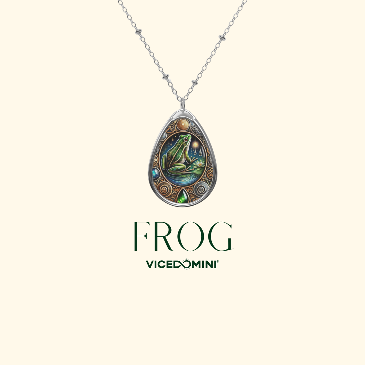 A Vicedomini 'Frog' pendant necklace featuring a silver teardrop-shaped frame with a detailed frog illustration and iridescent green and blue accents, suspended from a silver chain on a light cream background with the brand name and product name printed below.